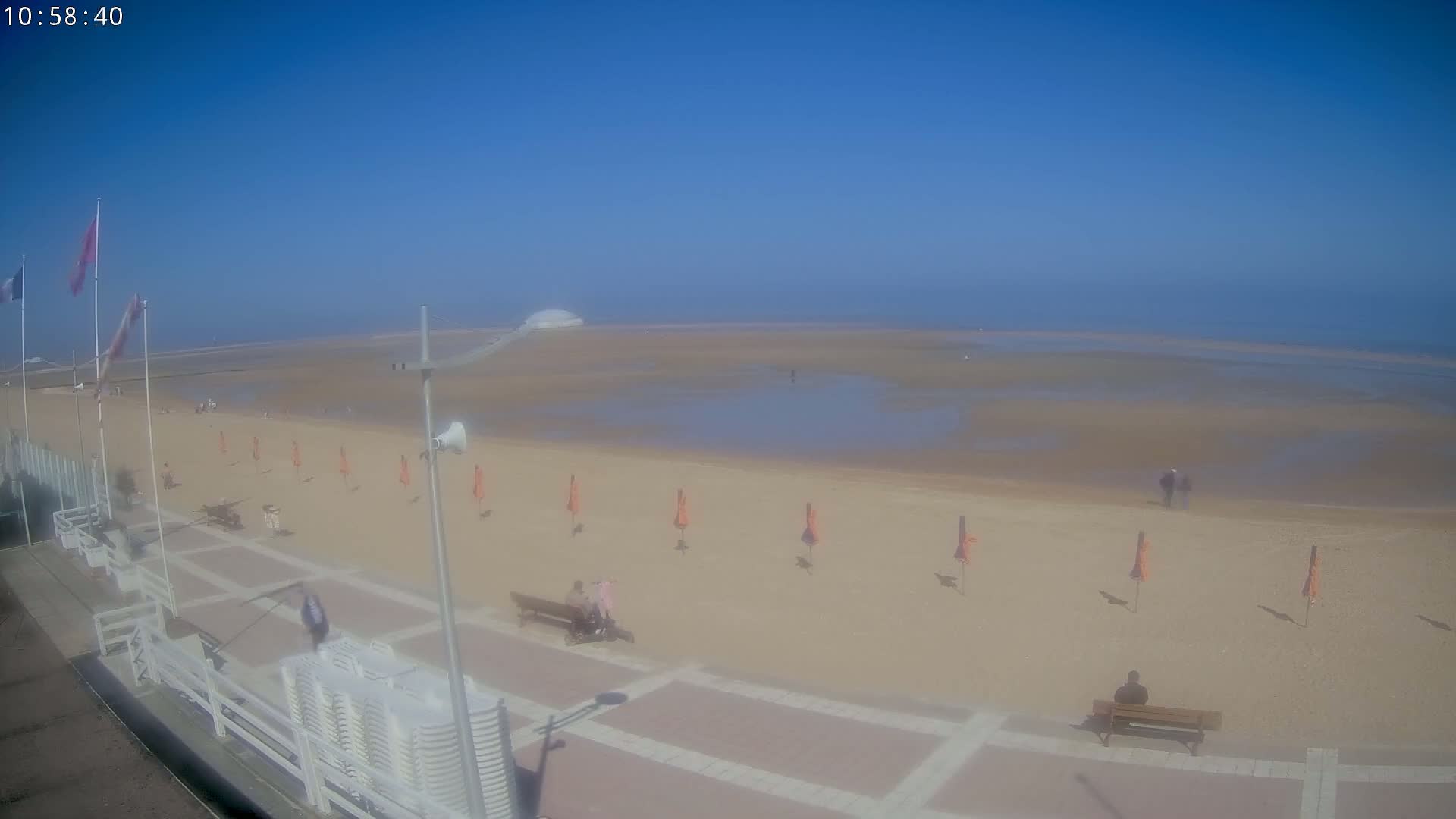 A mostly sunny beach scene shows a wide expanse of wet sand leading to a calm ocean under a clear blue sky.
