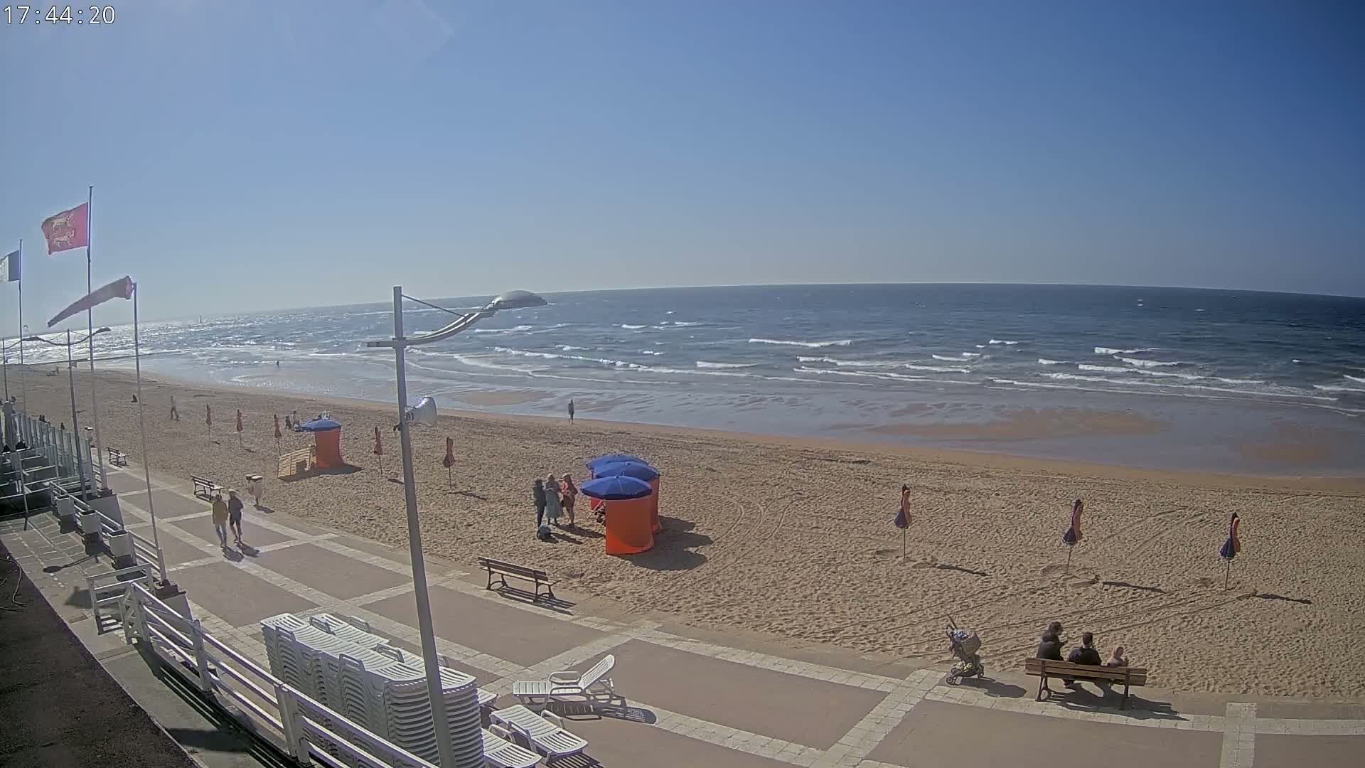 A sandy beach with a boardwalk and several people, under a clear blue sky with a moderately wavy ocean.