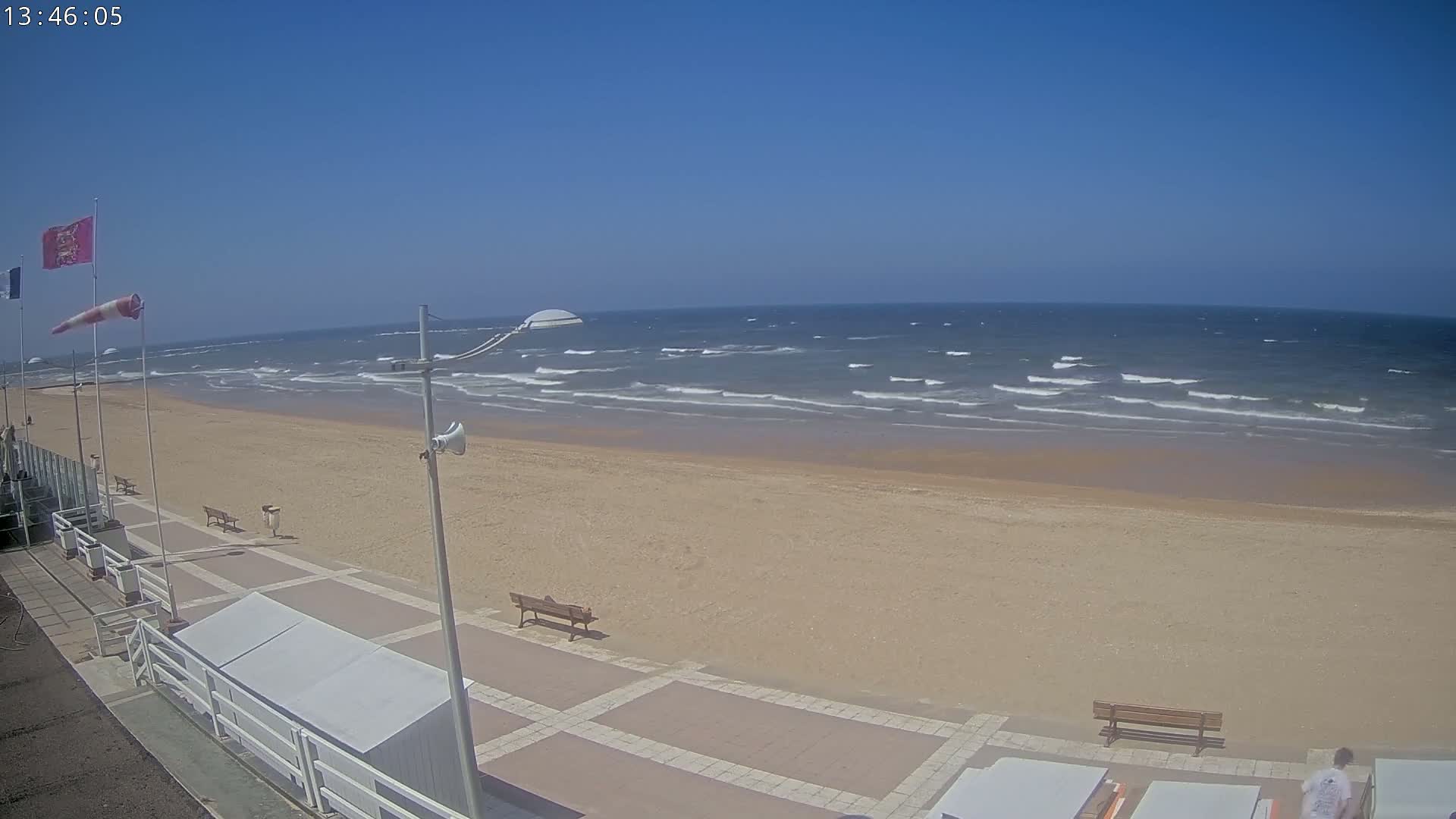 A wide sandy beach with small waves under a clear blue sky is shown, with a paved walkway and benches visible in the foreground.