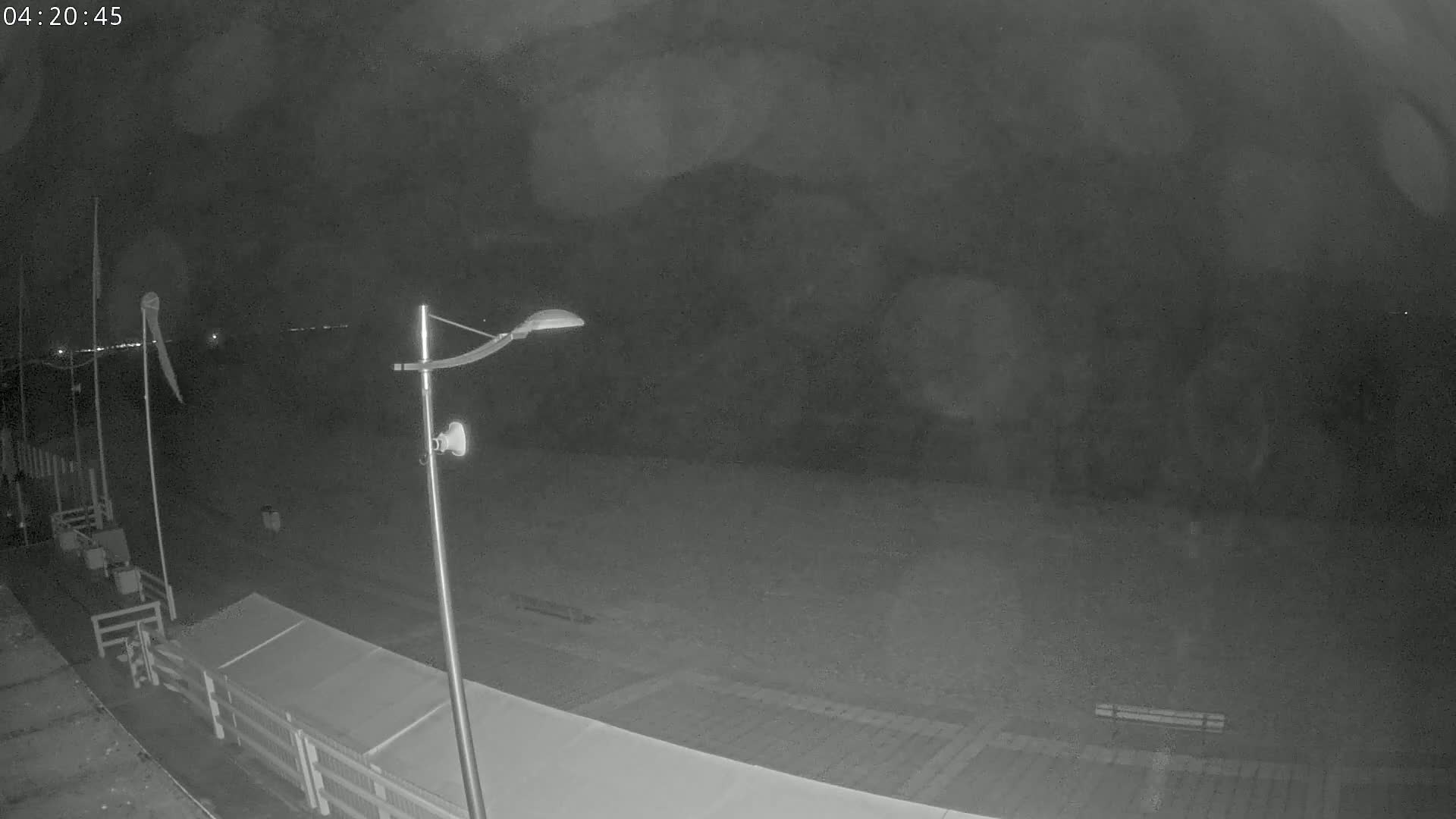 A nighttime, low-light view of a mostly empty beach area with a lamppost, some structures, and flags.