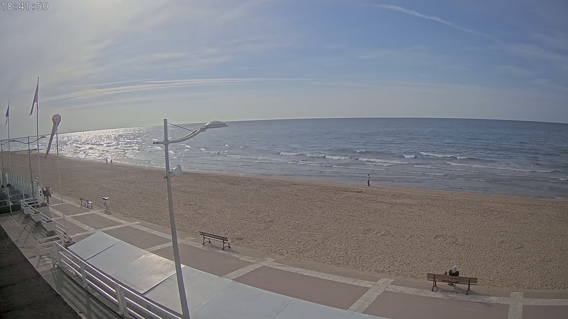 A sandy beach with a few people and benches, overlooking a calm ocean under a mostly clear sky.