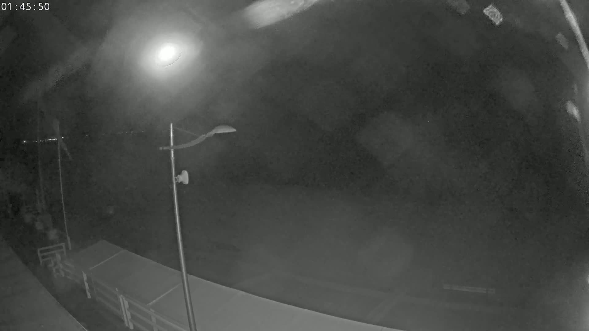 A nighttime, low-light view shows a lamppost on a deck overlooking a dark body of water under a cloudy sky.