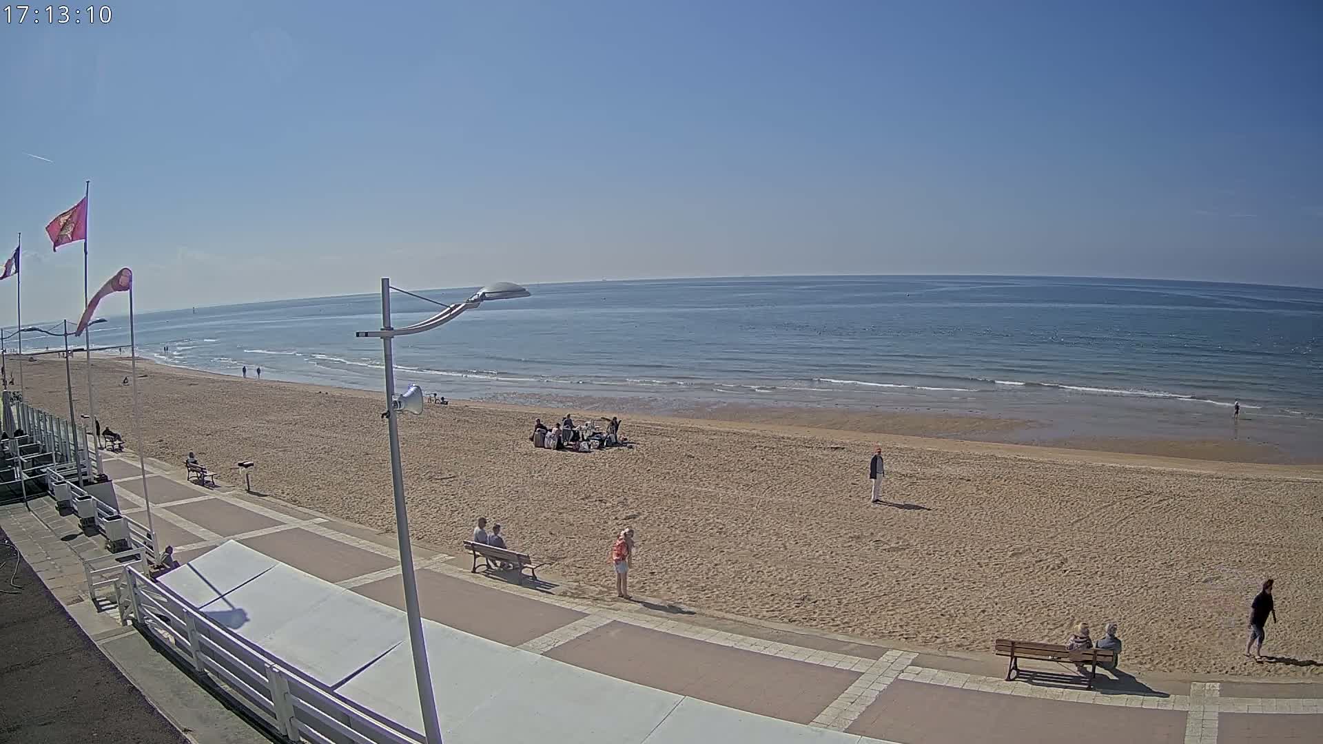 A sandy beach with a few people scattered across it and along a paved walkway, under a clear blue sky.