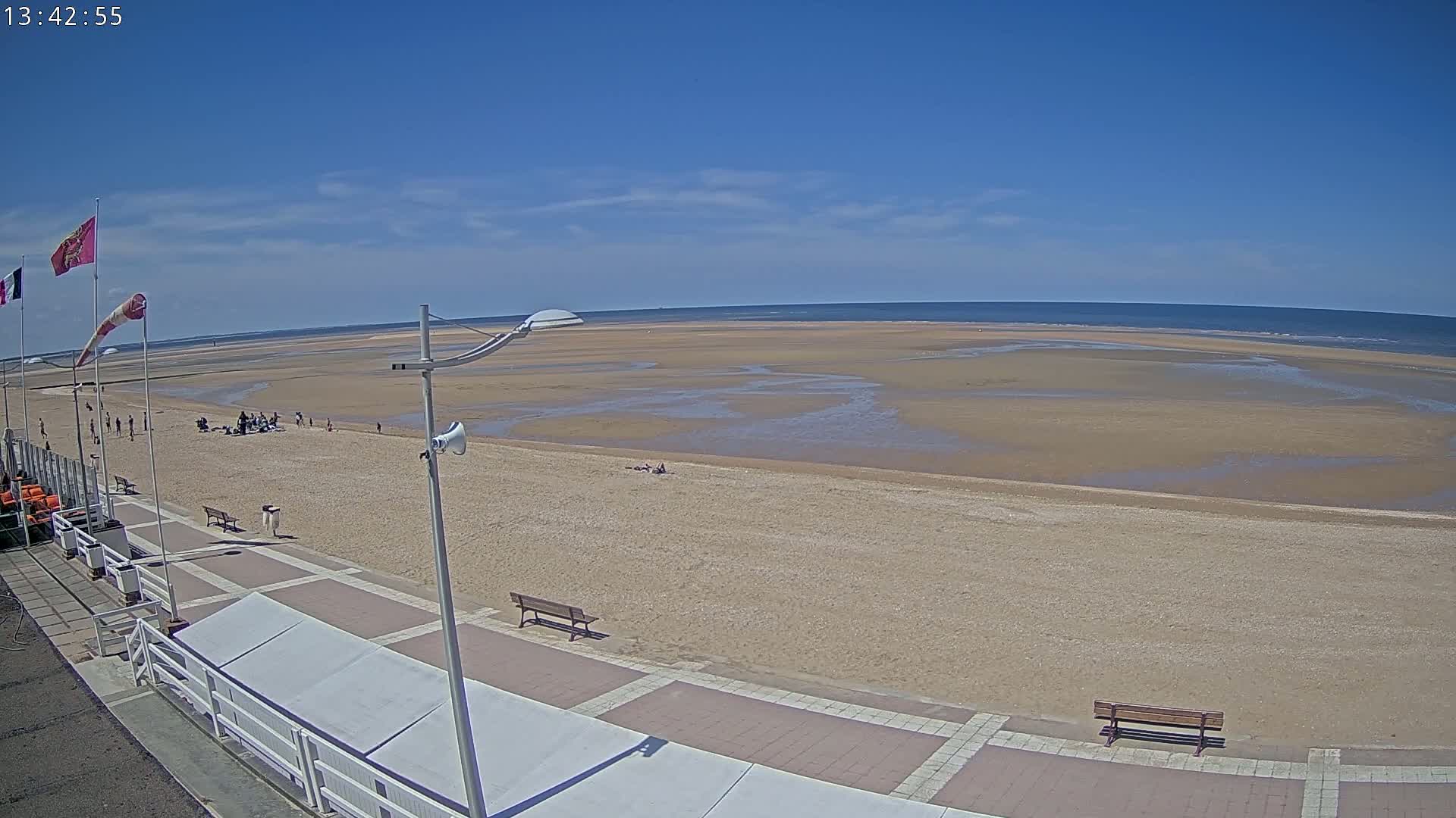 A sandy beach stretches to a calm ocean under a clear blue sky, with a boardwalk and a few people visible.