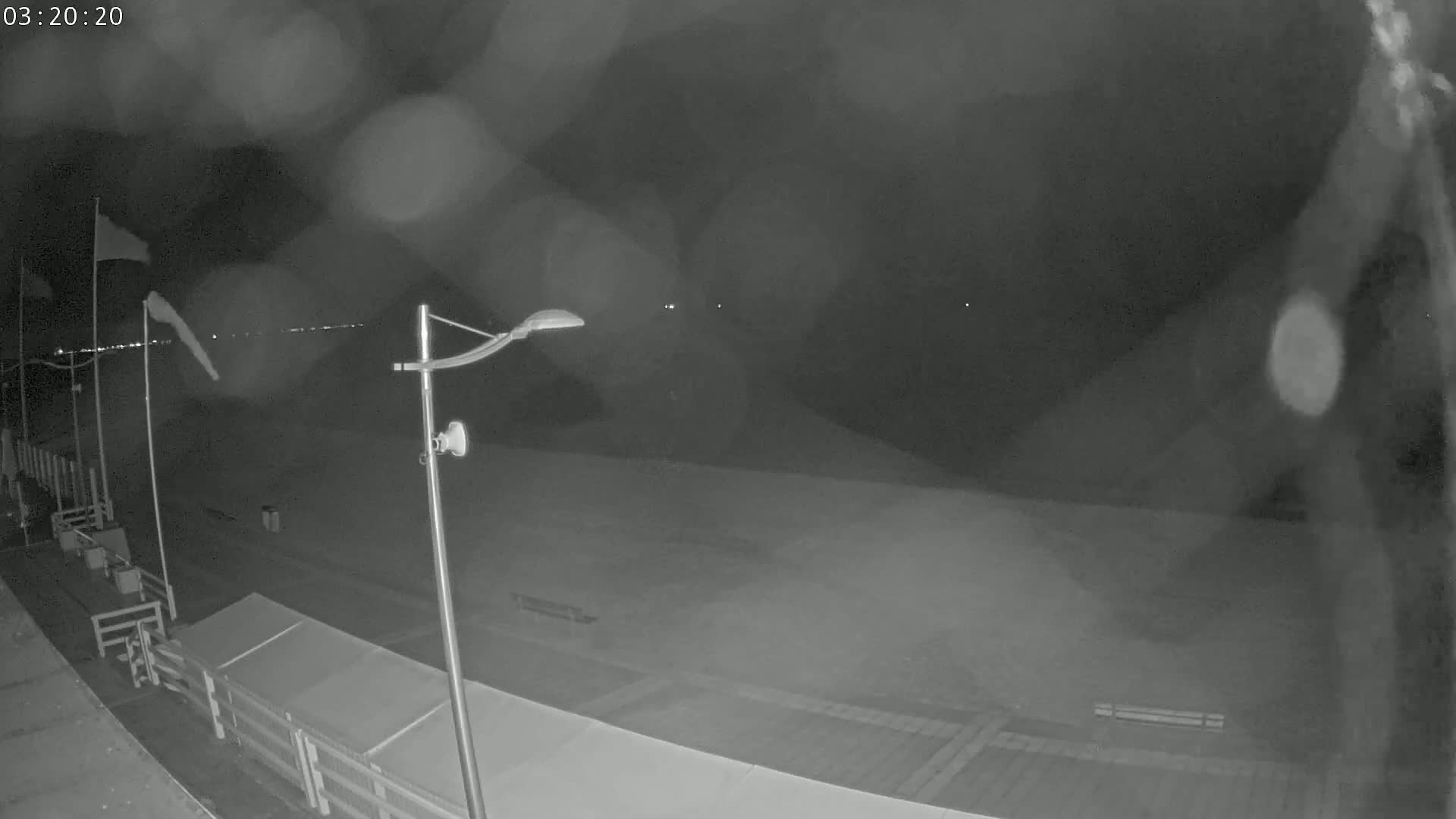 A nighttime, blurry view of a mostly empty beach boardwalk with a streetlight and flags in the wind under a dark, possibly rainy sky.