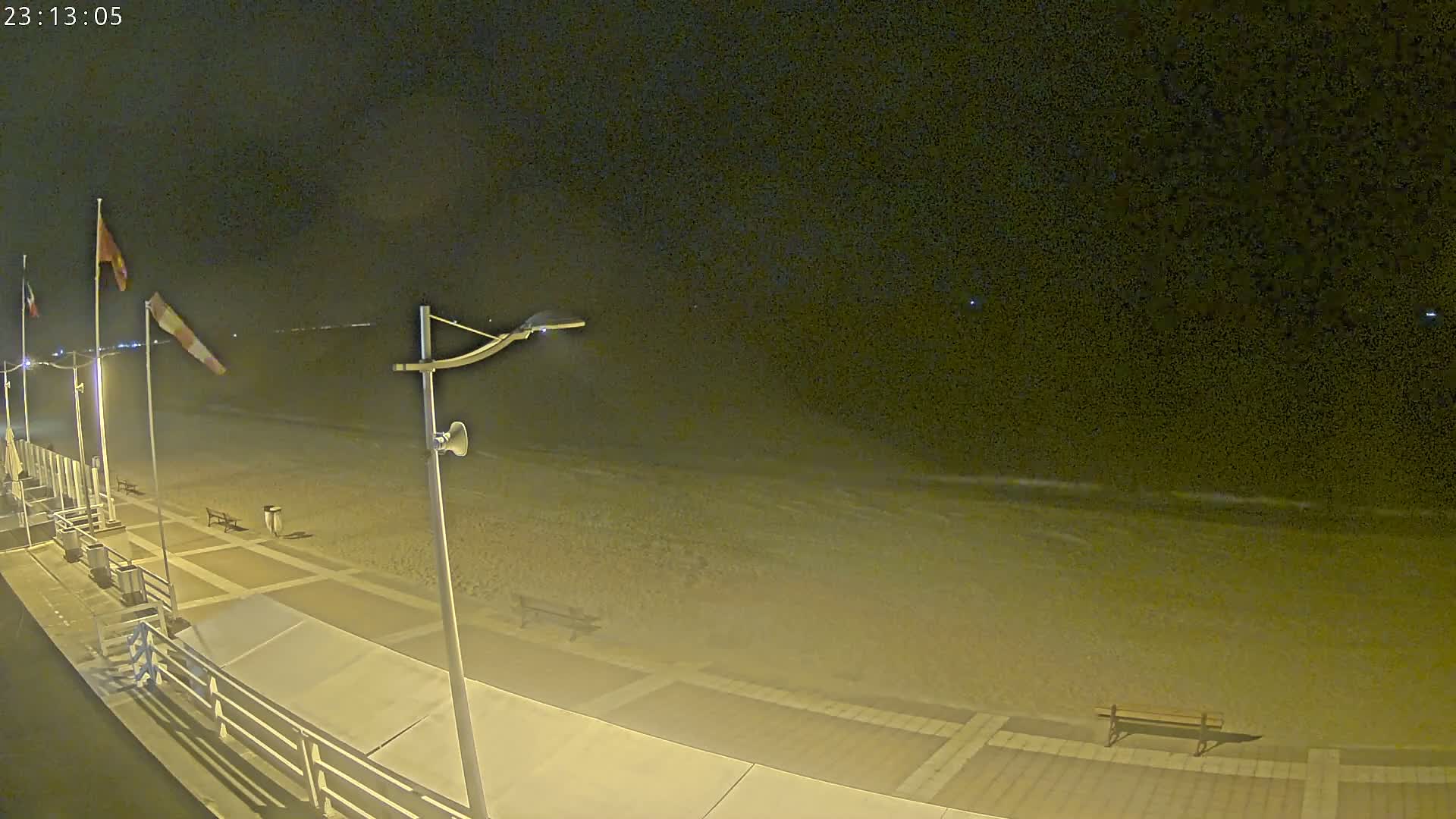 A nighttime view of a sandy beach with gentle waves under a dark sky, showing a boardwalk with benches and flags on poles.