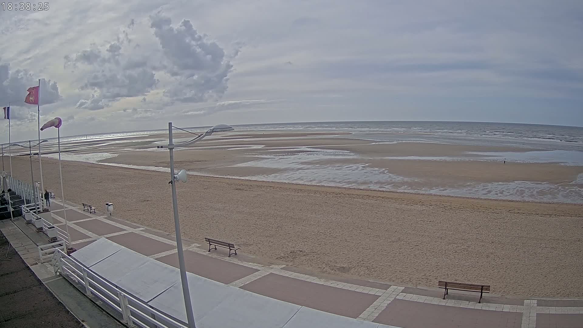 A mostly cloudy day at a wide sandy beach with a boardwalk, benches, and flags, showing a mostly receding tide.