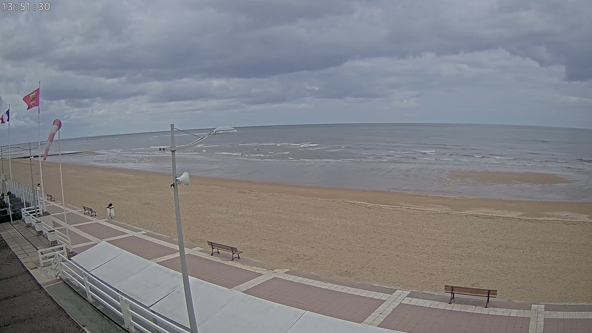 A mostly cloudy day shows a sandy beach with a paved walkway, benches, and flags, overlooking a calm sea with small waves.