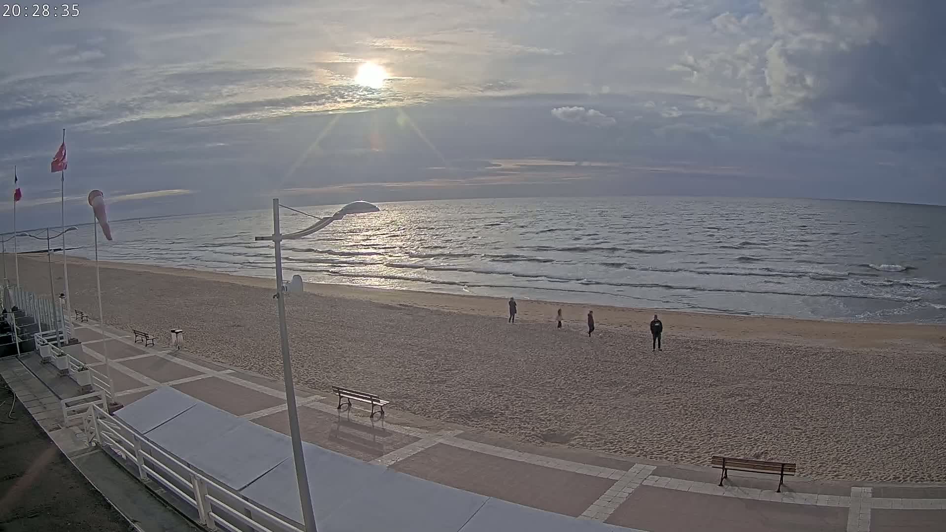 A mostly sunny day shows a sandy beach with a few people walking along the shoreline, and a paved walkway with benches leading to the ocean under a partly cloudy sky.