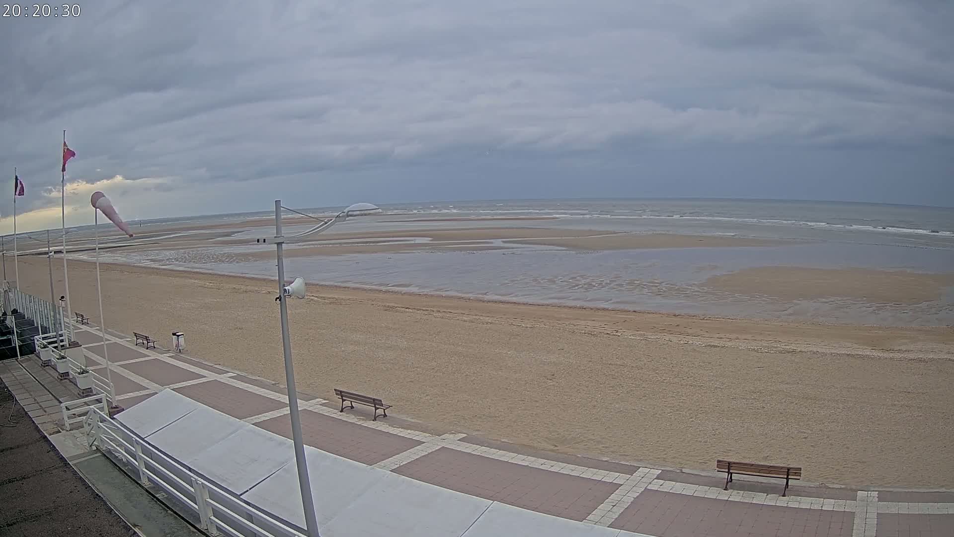 A mostly cloudy day shows a wide sandy beach with a paved walkway and benches, overlooking a calm sea at low tide.