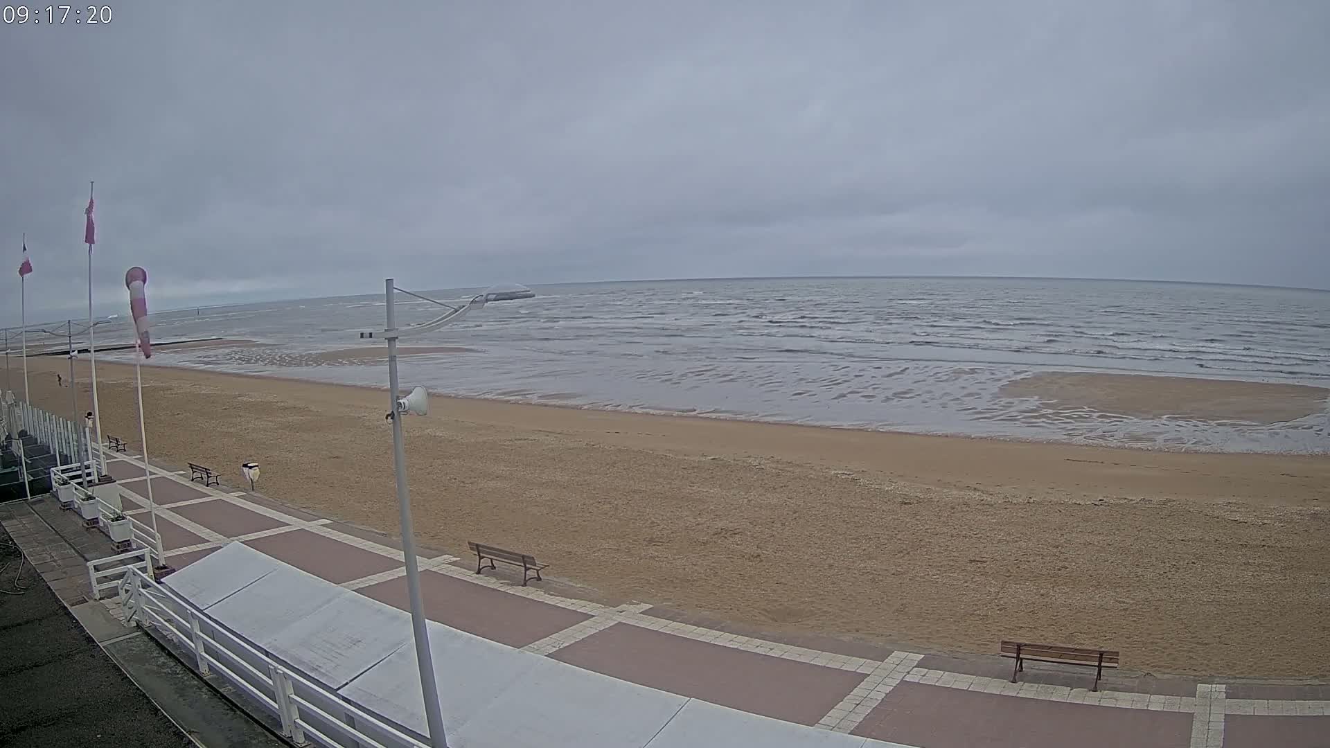 A sandy beach with a paved walkway and benches stretches to a grey, overcast sea with small waves.