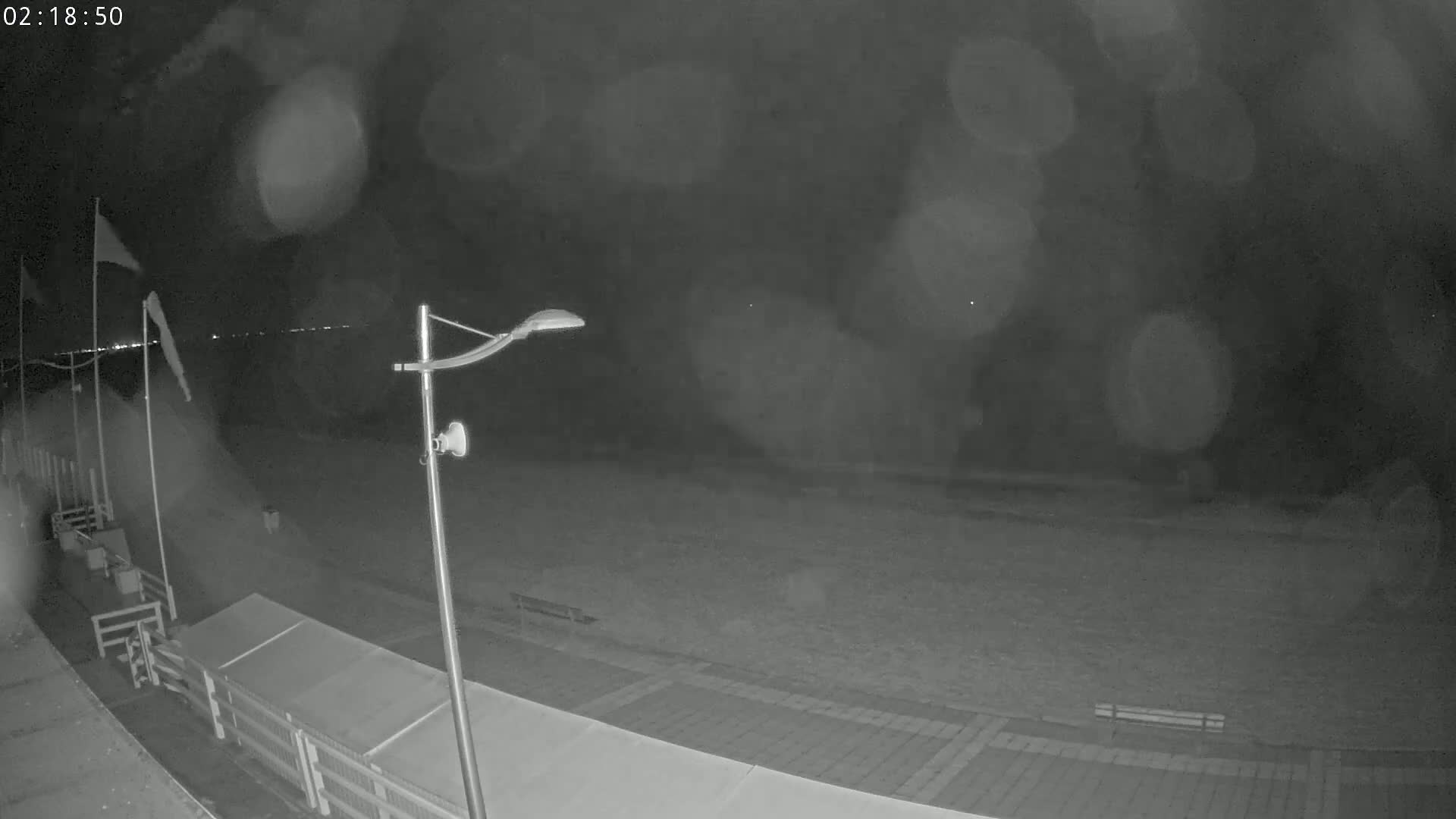 A nighttime, hazy view shows a mostly empty paved area near a body of water, with a lamppost and flags visible in the foreground.