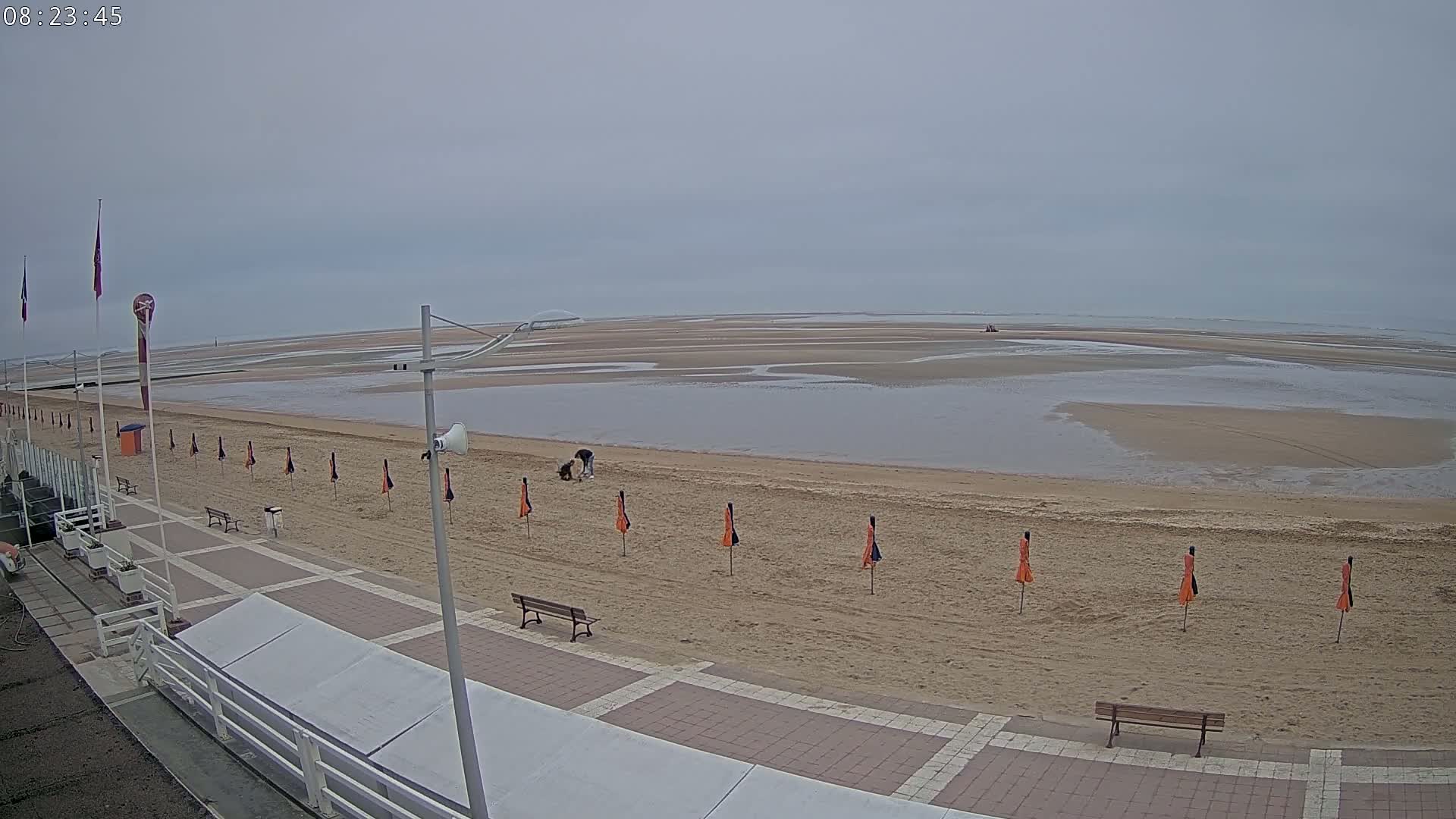 A mostly empty beach with a few people near the water's edge under an overcast sky, featuring a paved walkway with benches and folded beach umbrellas.