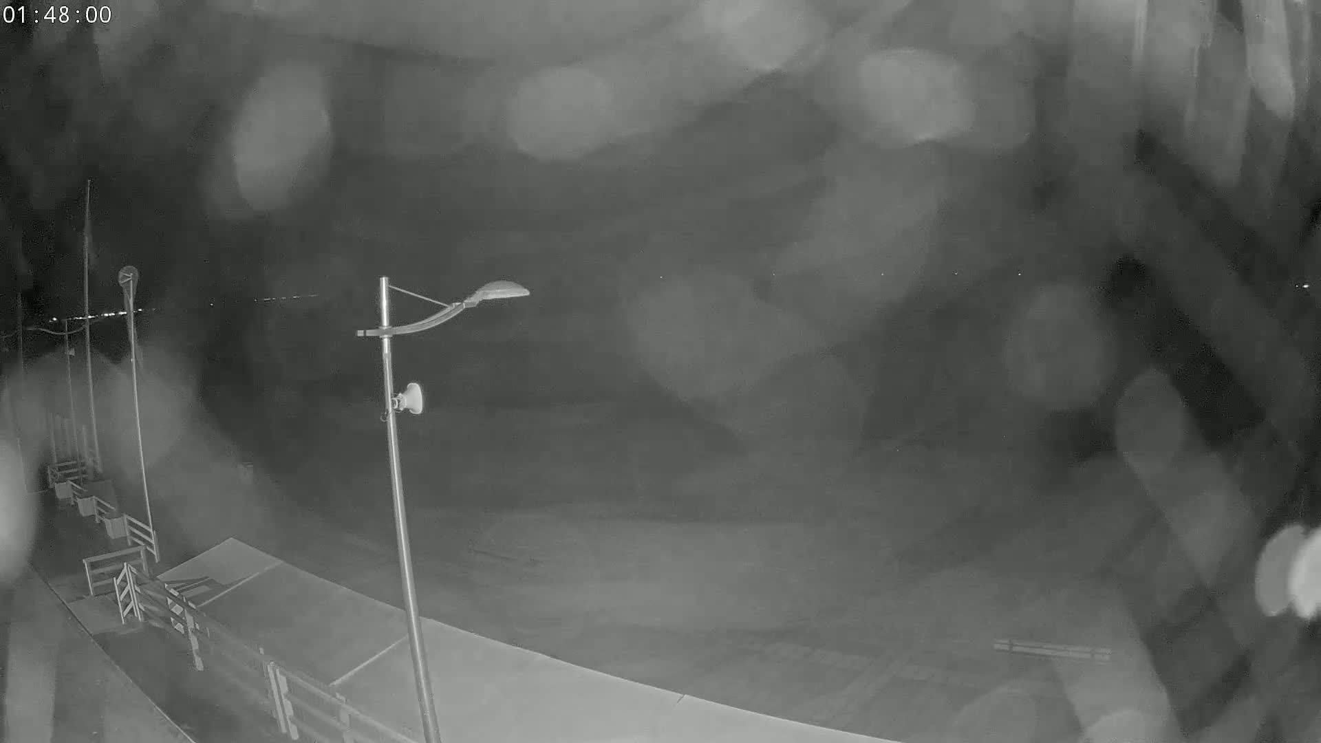 A nighttime, blurry, low-light view of a mostly empty dock or pier, with a lamppost and light rain or mist.