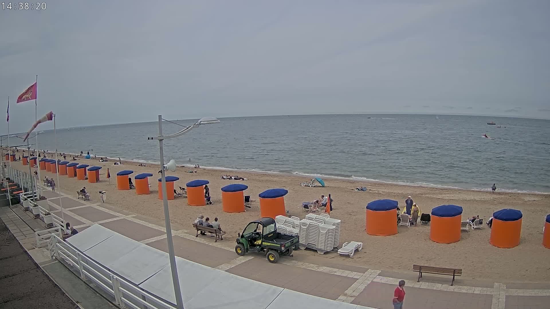 A sandy beach with orange and blue beach huts, a few people sunbathing, a small utility vehicle, and a calm, overcast sea.