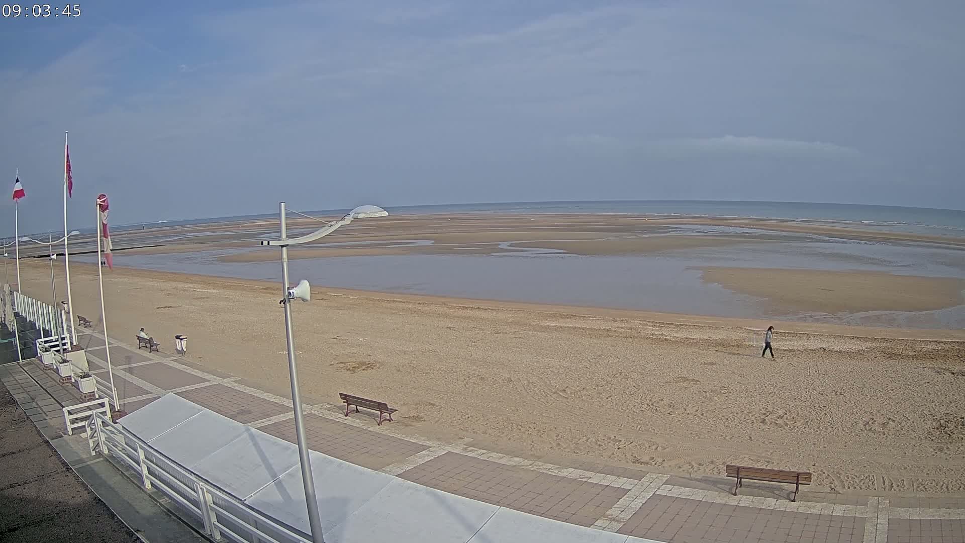 A mostly sunny day at a beach shows a wide expanse of sand at low tide, a paved walkway with benches, and a few people walking.