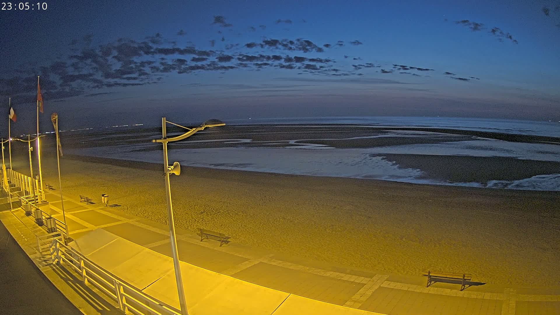 A mostly empty, sandy beach at night under a twilight sky with a few clouds is seen from a high vantage point, showing a paved walkway with benches and lampposts.