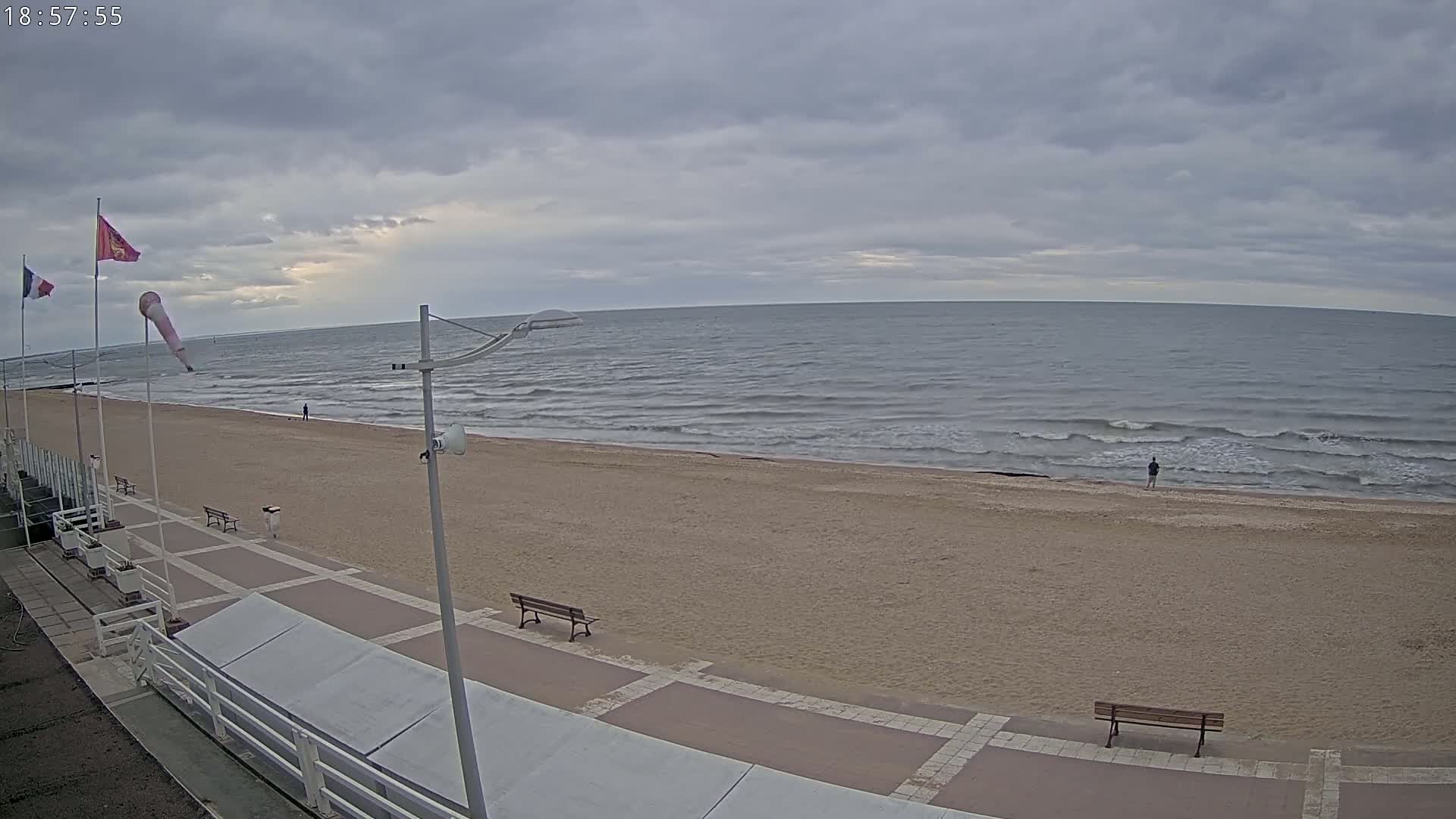 A sandy beach with a boardwalk and benches stretches along a wavy ocean under a cloudy sky.