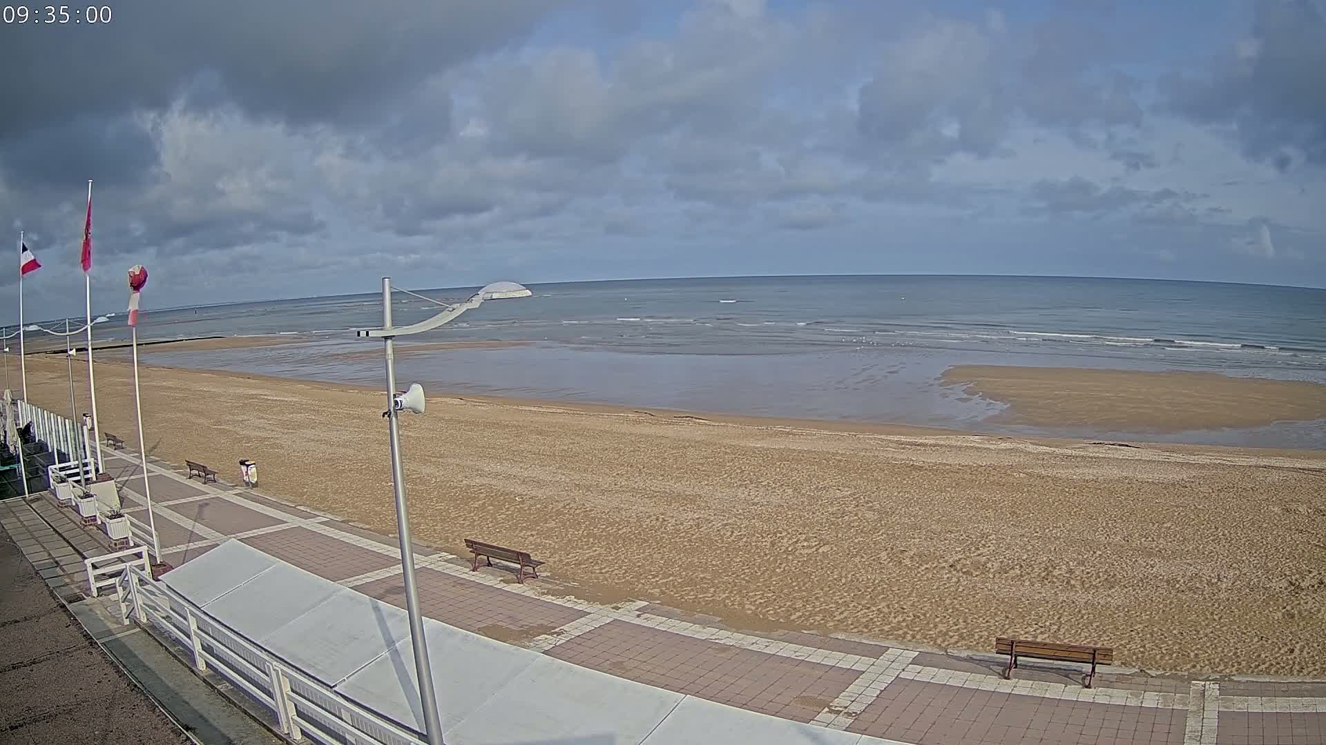A mostly cloudy day reveals a wide, sandy beach with a paved walkway and benches, overlooking a calm ocean with small waves.