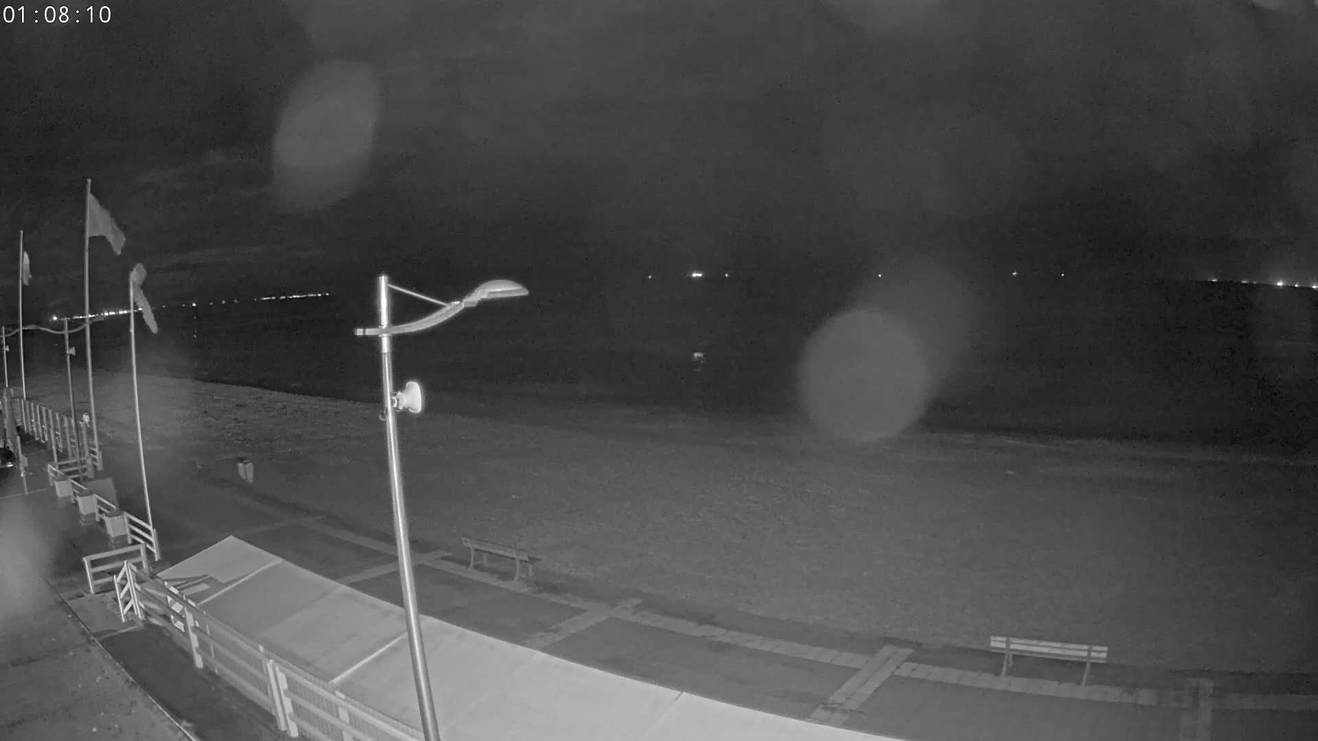 A nighttime, rainy view of a mostly empty beach with a boardwalk, a few flags, a lamppost, and a bench.
