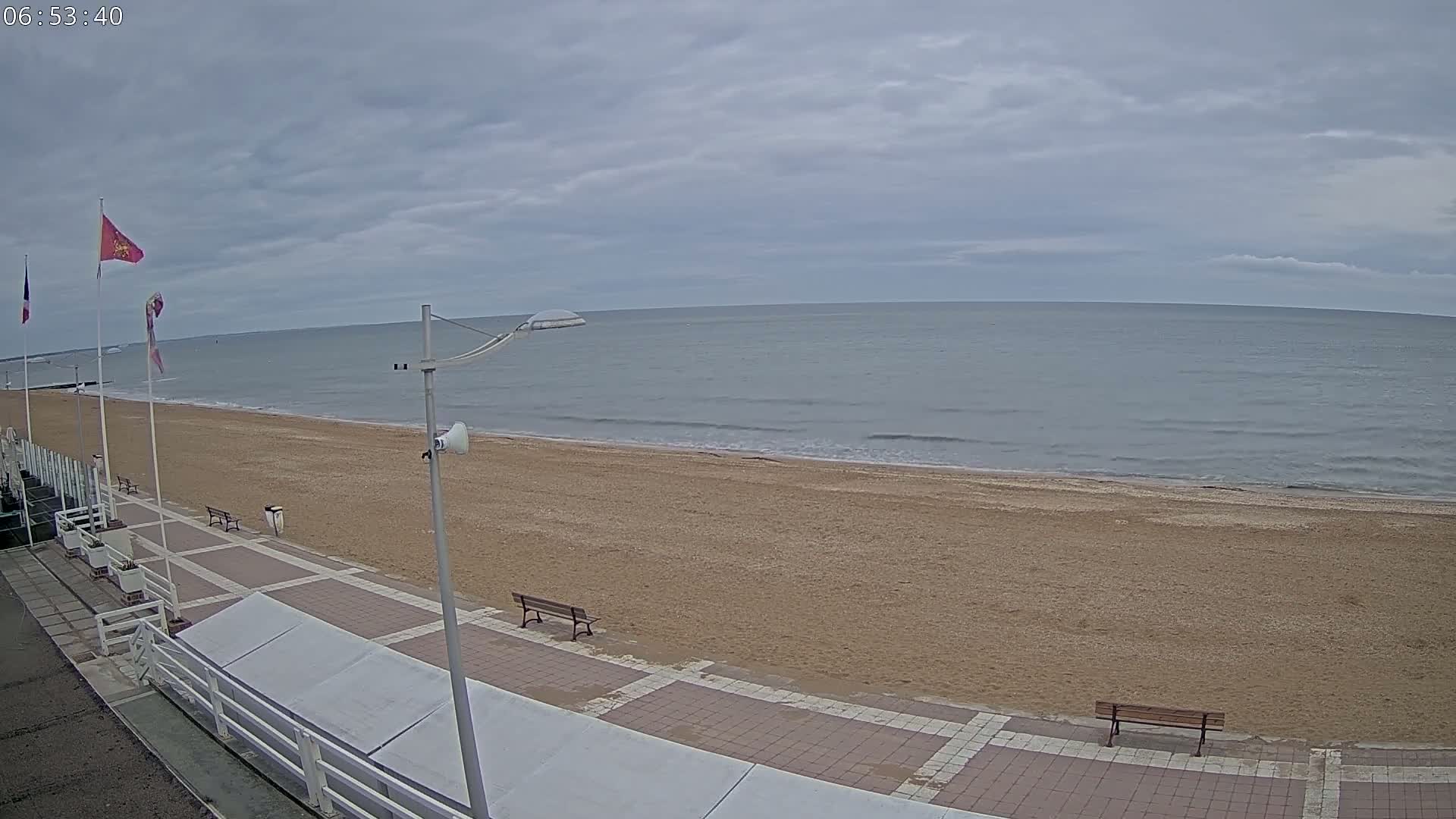 A sandy beach with calm, gray water under an overcast sky is seen from a boardwalk with two benches.