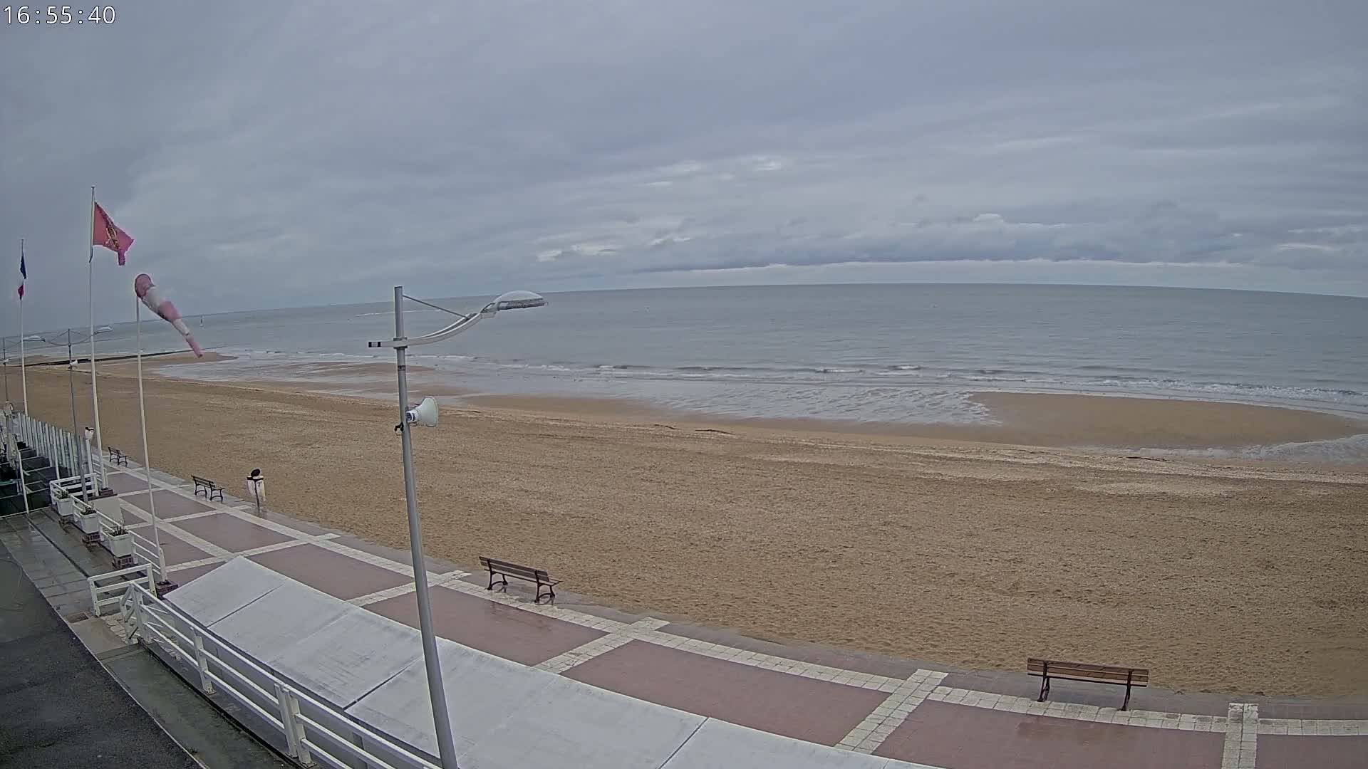 A mostly cloudy day shows a sandy beach with a paved walkway and benches, overlooking a calm sea with small waves.