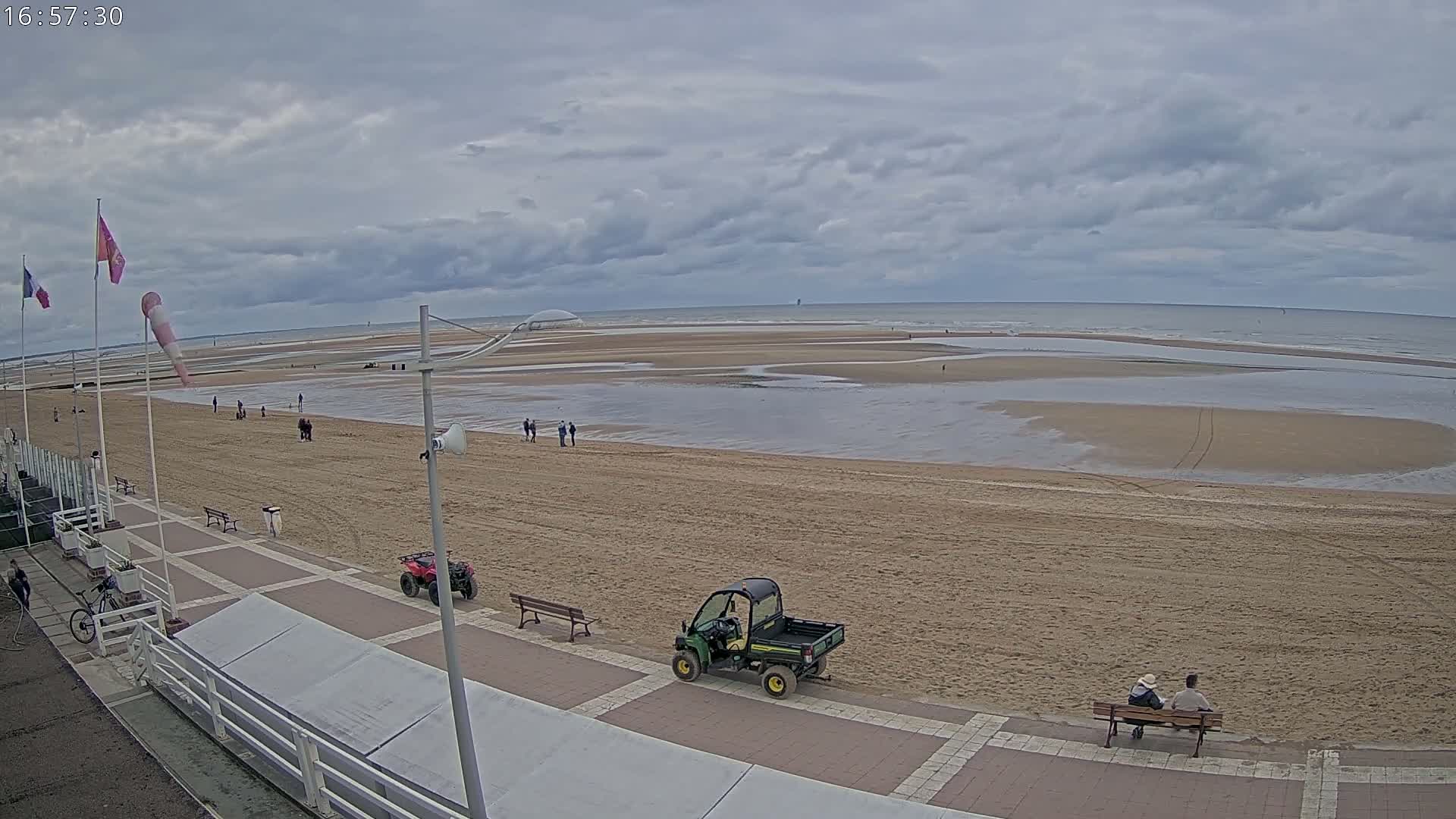A sandy beach with a few people and small vehicles is seen from an elevated vantage point under a cloudy sky, with calm ocean waves in the background.