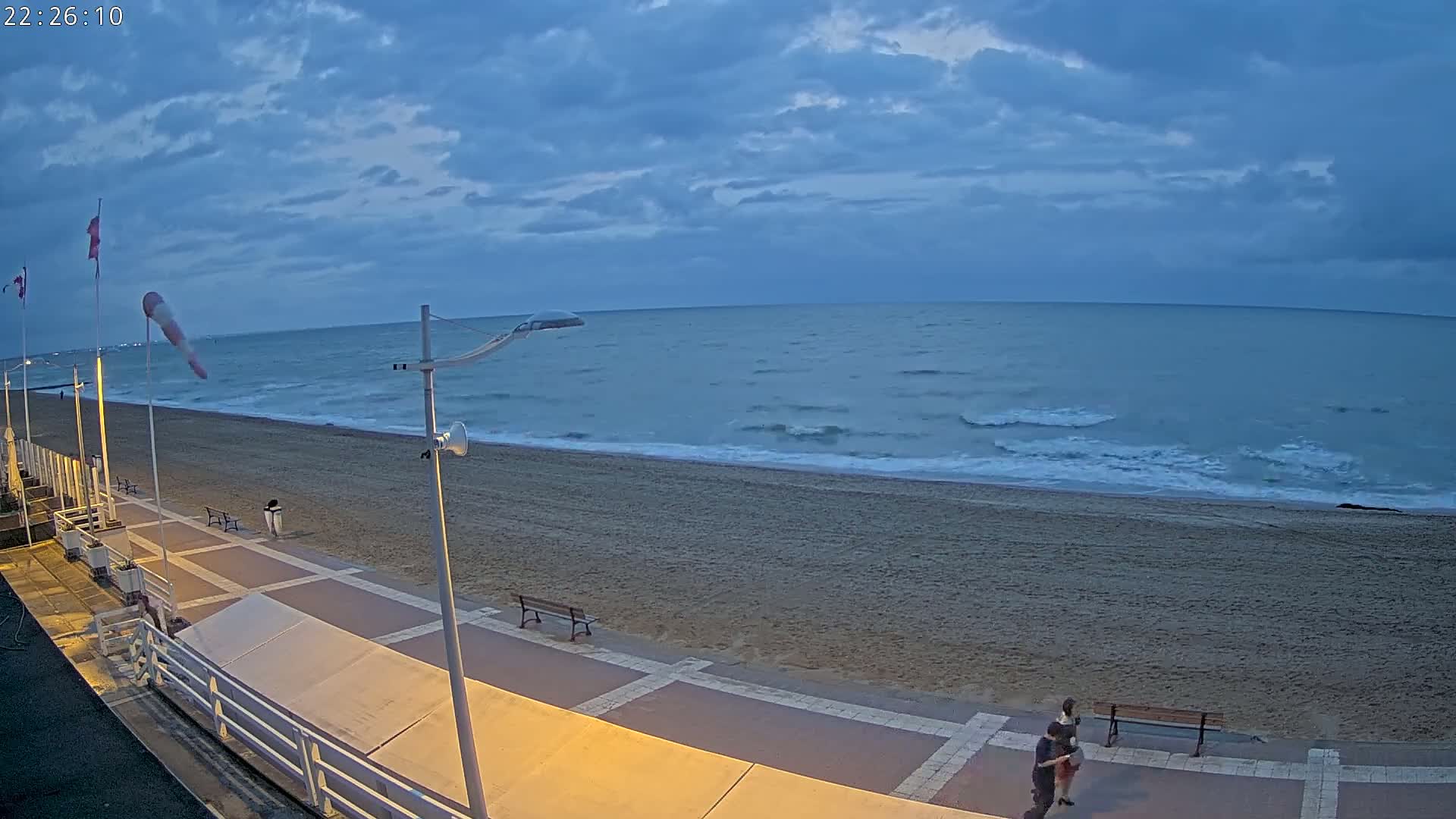 A mostly cloudy twilight sky overlooks a sandy beach with a paved walkway, streetlights, and two people walking near a bench.