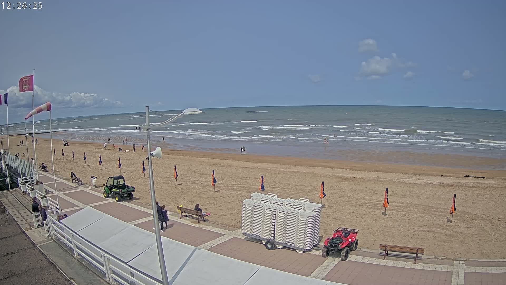 A mostly sunny day at a beach shows a wide sandy shore with small waves, several people, beach umbrellas, and utility vehicles parked on the boardwalk.