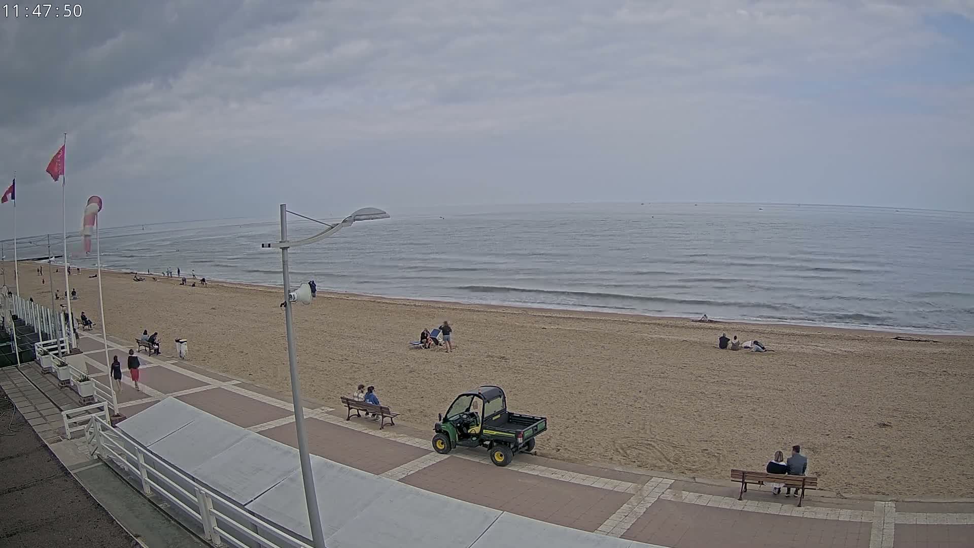 A sandy beach with a few people scattered on it and along a paved walkway, under an overcast sky, is viewed from above.