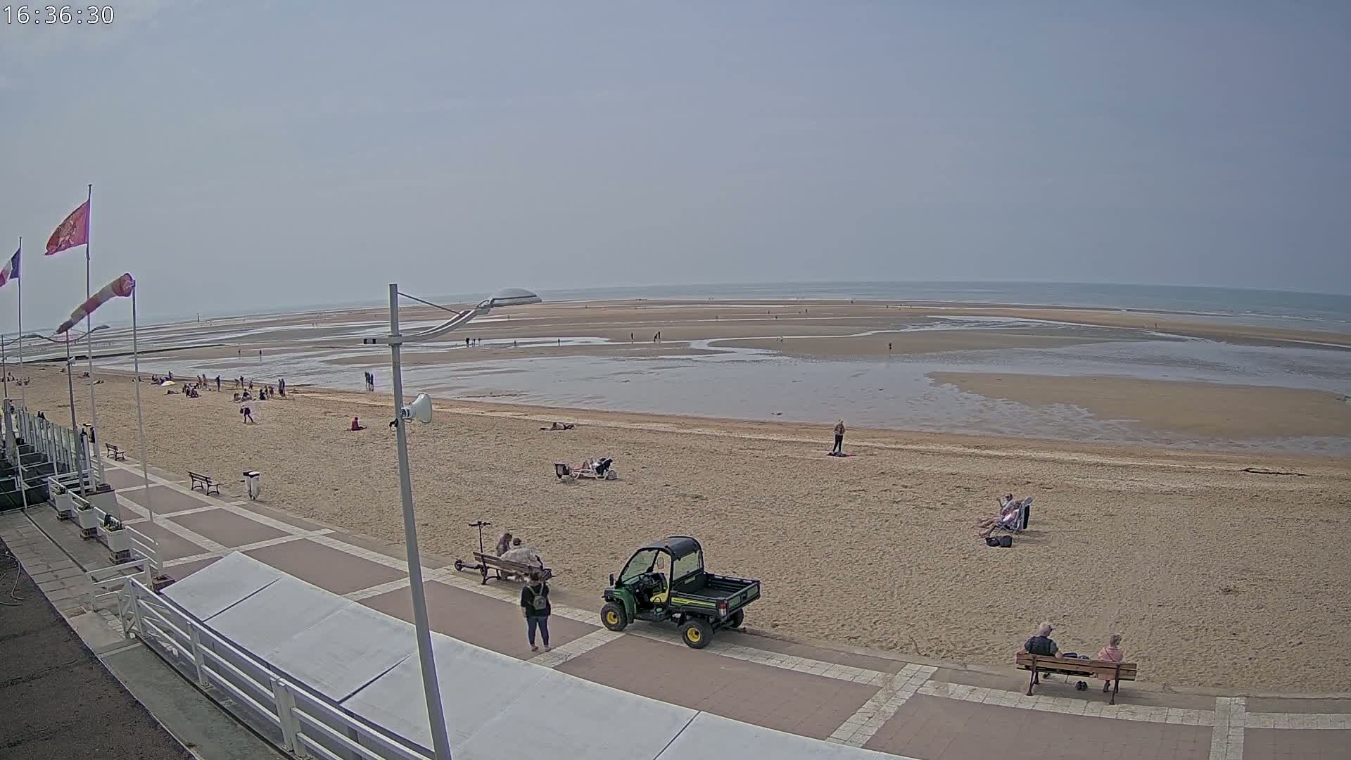 A sandy beach with scattered people, a utility vehicle, and a boardwalk stretches to a calm, shallow ocean under a hazy sky.
