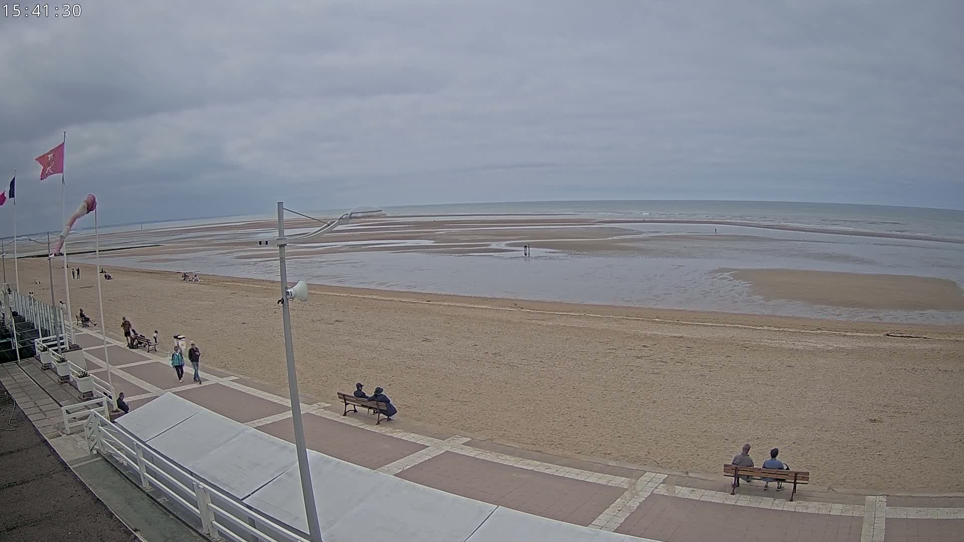 A wide sandy beach stretches to a calm sea under a cloudy sky, with a few people strolling along a paved walkway and sitting on benches.