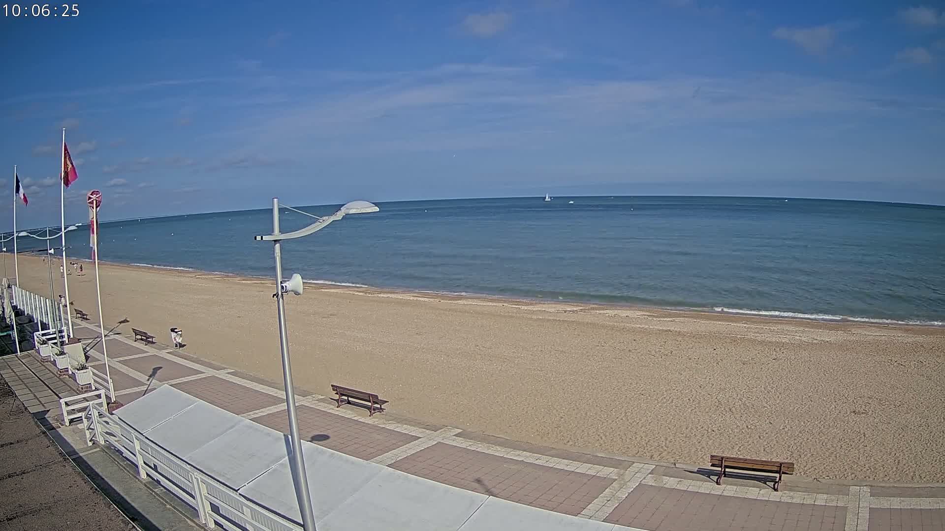 A sandy beach with a paved walkway and benches stretches along a calm, blue ocean under a clear, sunny sky.