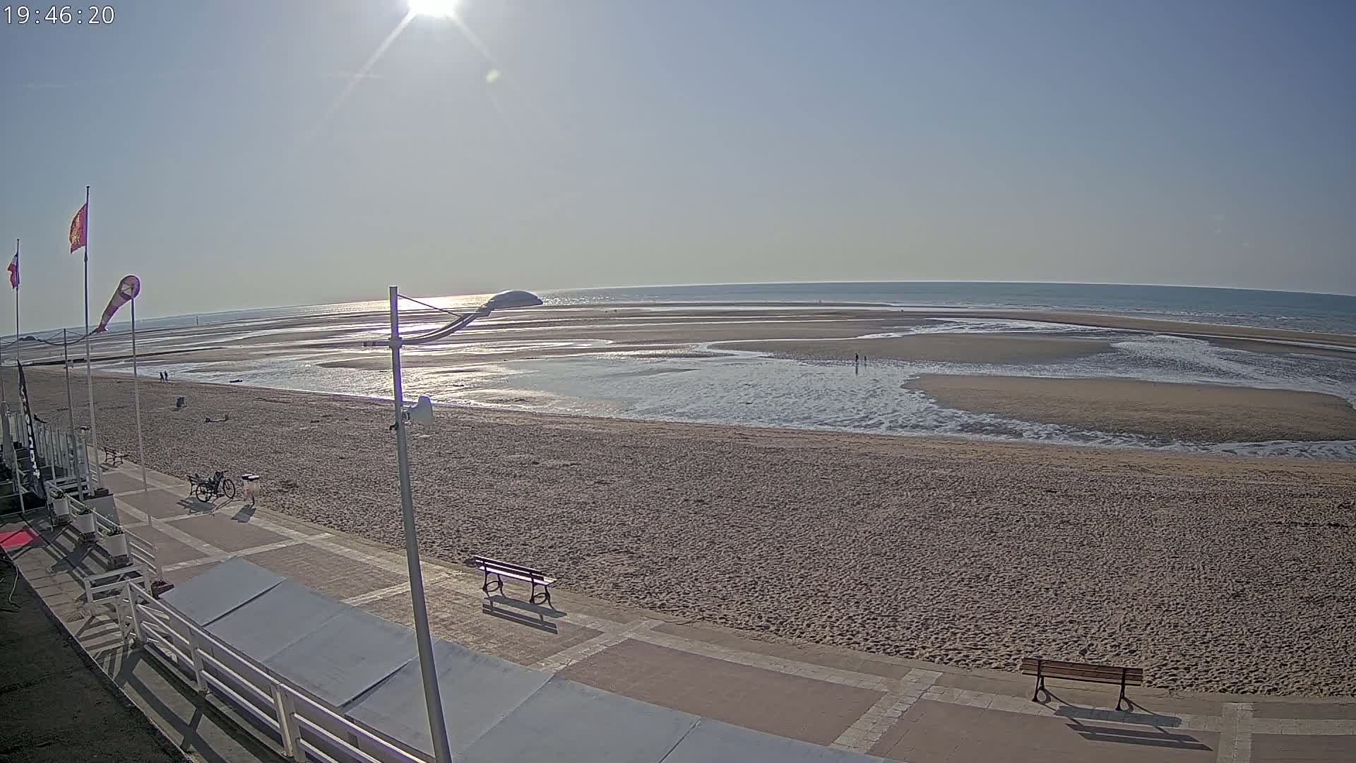 A sunny beach scene shows a wide expanse of sand and shallow water receding from the ocean, with a paved walkway and benches along the shoreline.