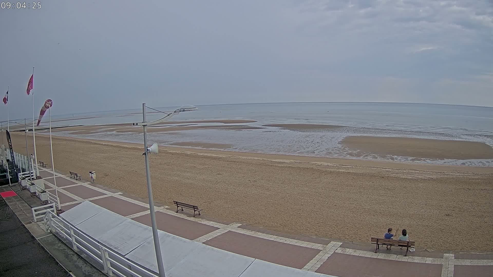 A sandy beach with a calm sea under an overcast sky, and two people sitting on a bench overlooking the water.