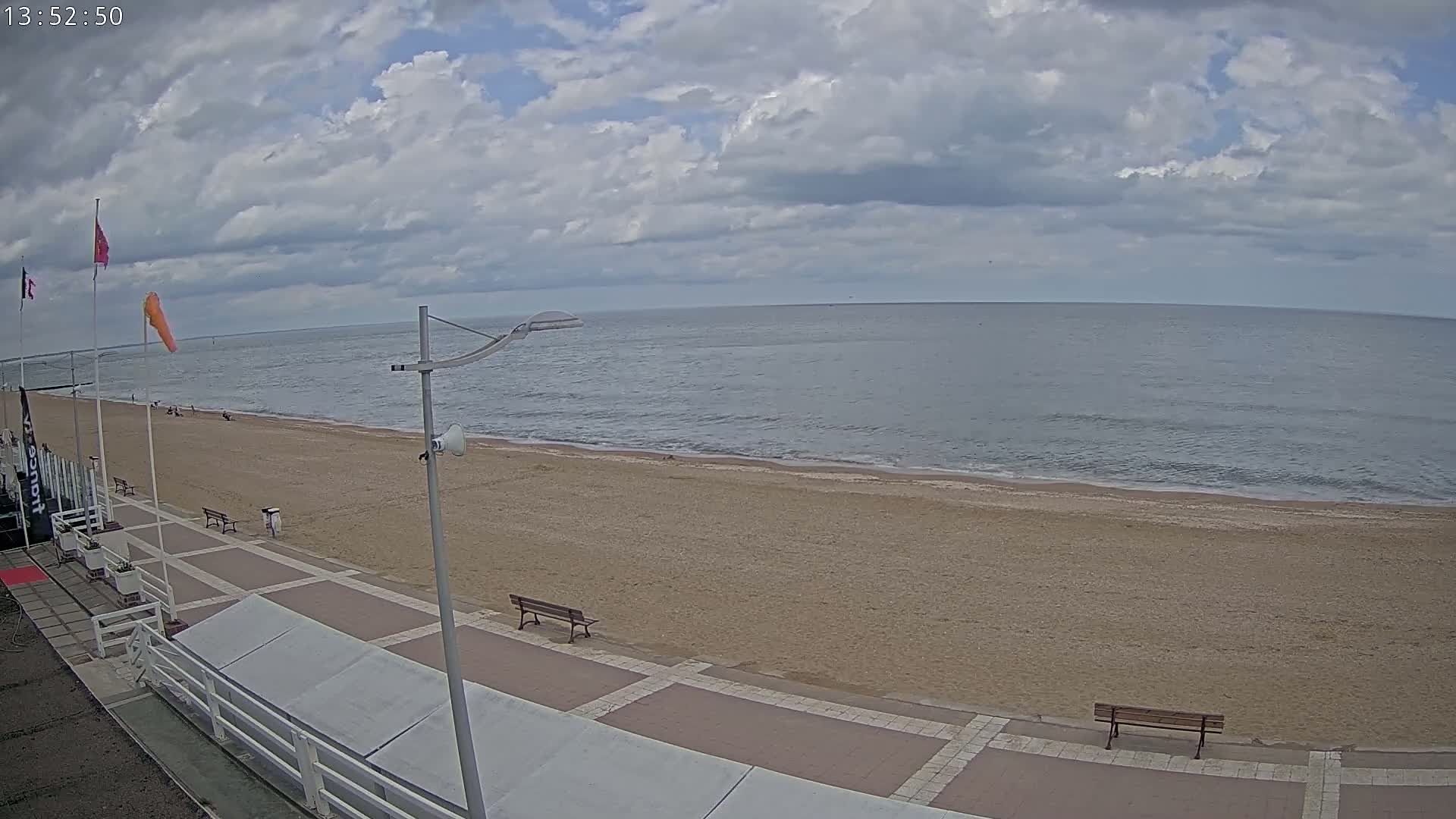 A mostly cloudy day at a sandy beach with calm ocean waters, showing a boardwalk with benches and a few people in the distance.