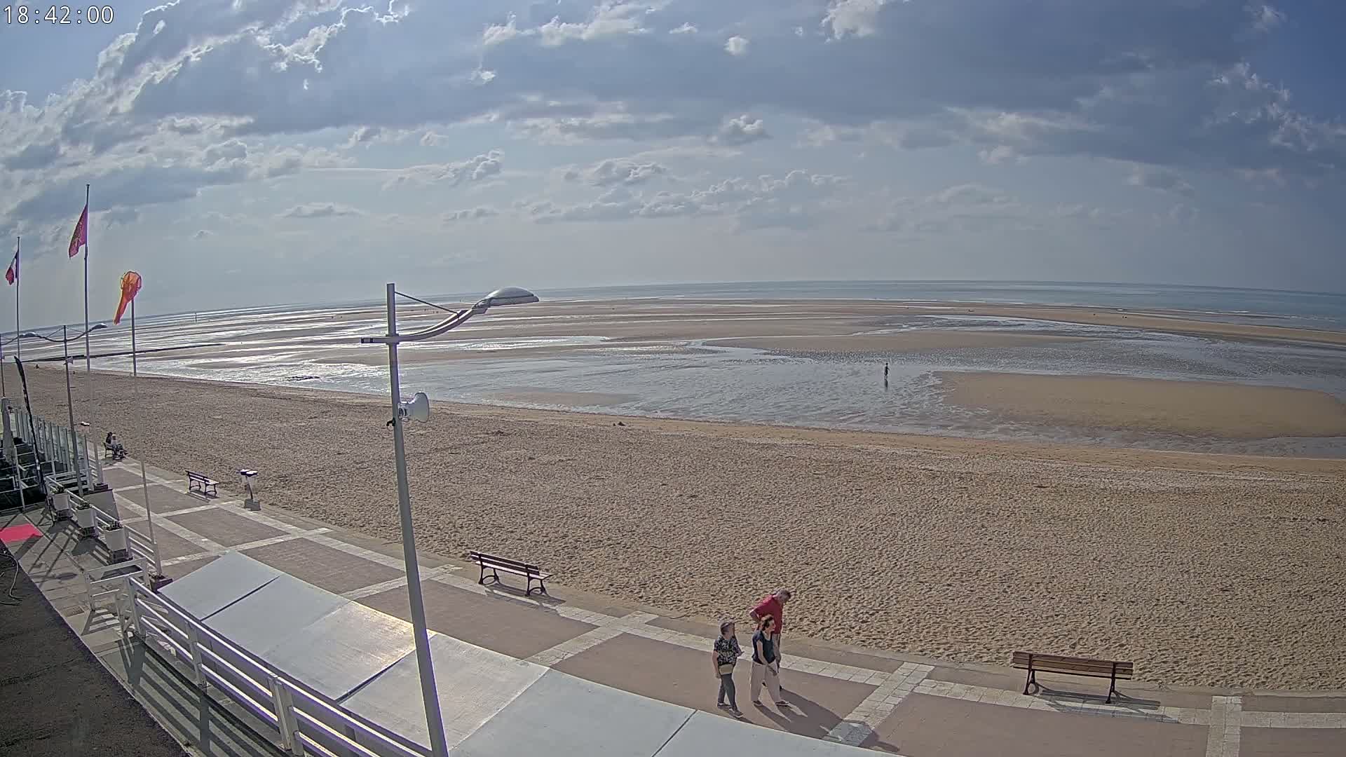 A wide sandy beach stretches to a calm ocean under a partly cloudy sky, with a paved walkway and a few people strolling nearby.
