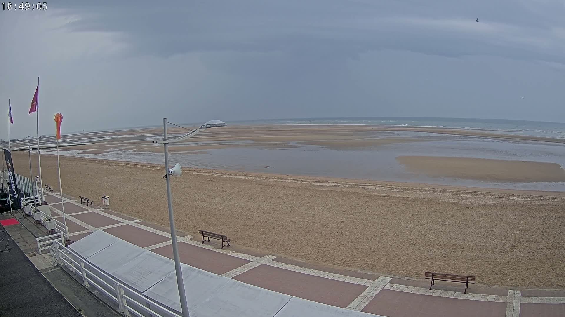 A mostly overcast day shows a wide sandy beach with calm water receding to reveal exposed sandbars, and a paved boardwalk with benches along the edge.