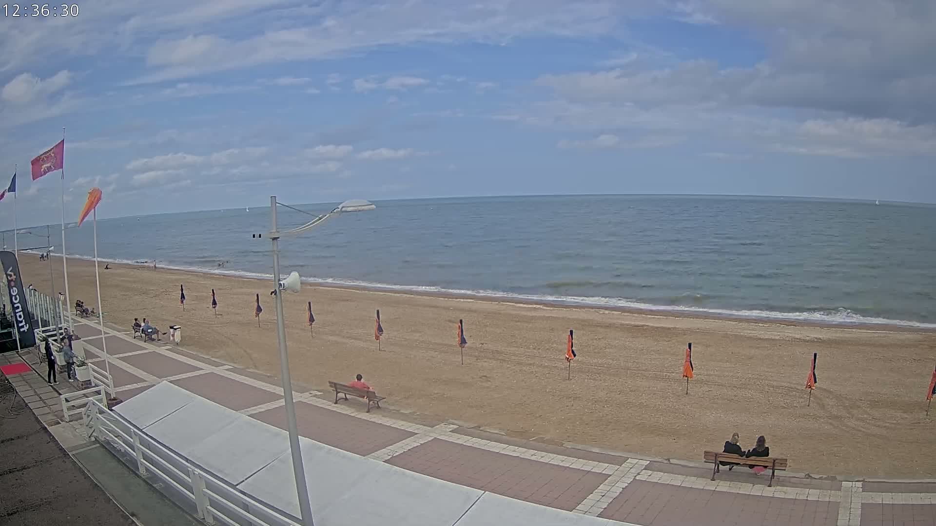 A sandy beach with a few people and orange umbrellas stretches along a calm sea under a partly cloudy sky.