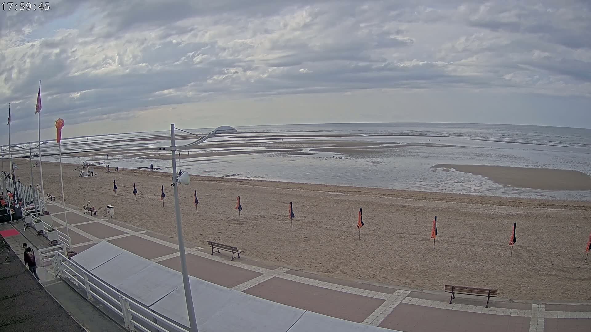 A mostly cloudy day at a beach shows a wide sandy shore with a few people, several closed umbrellas, and benches on a paved walkway near a building.
