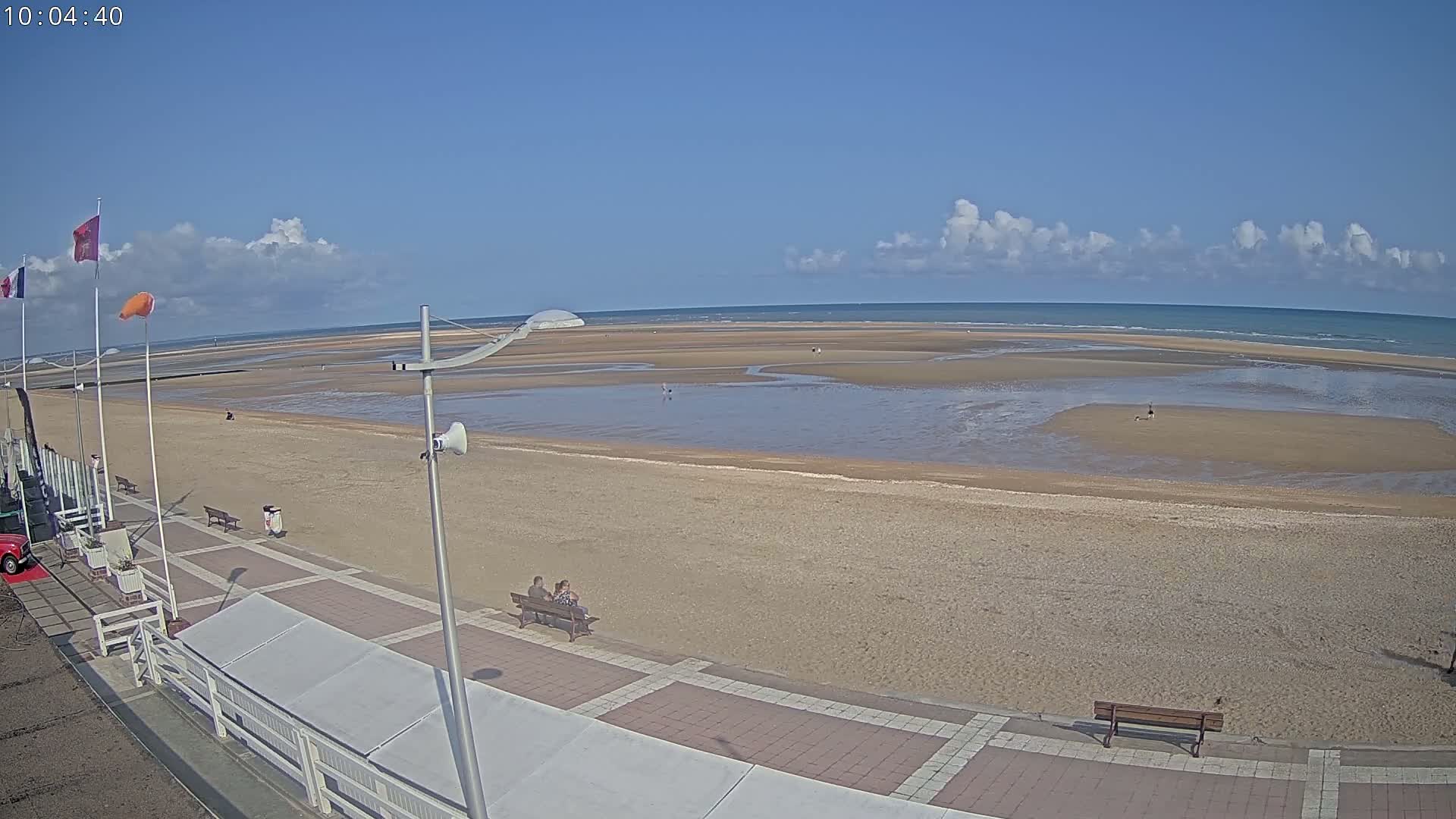 A mostly sunny beach scene shows a wide expanse of sand and shallow water under a partly cloudy sky.