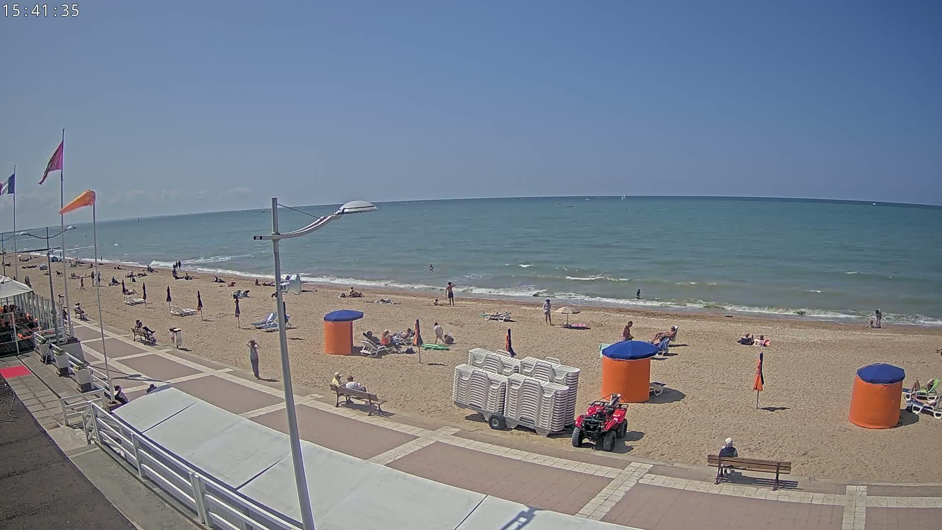 A sandy beach with scattered sunbathers and beach umbrellas is bordered by a paved walkway under a clear blue sky.