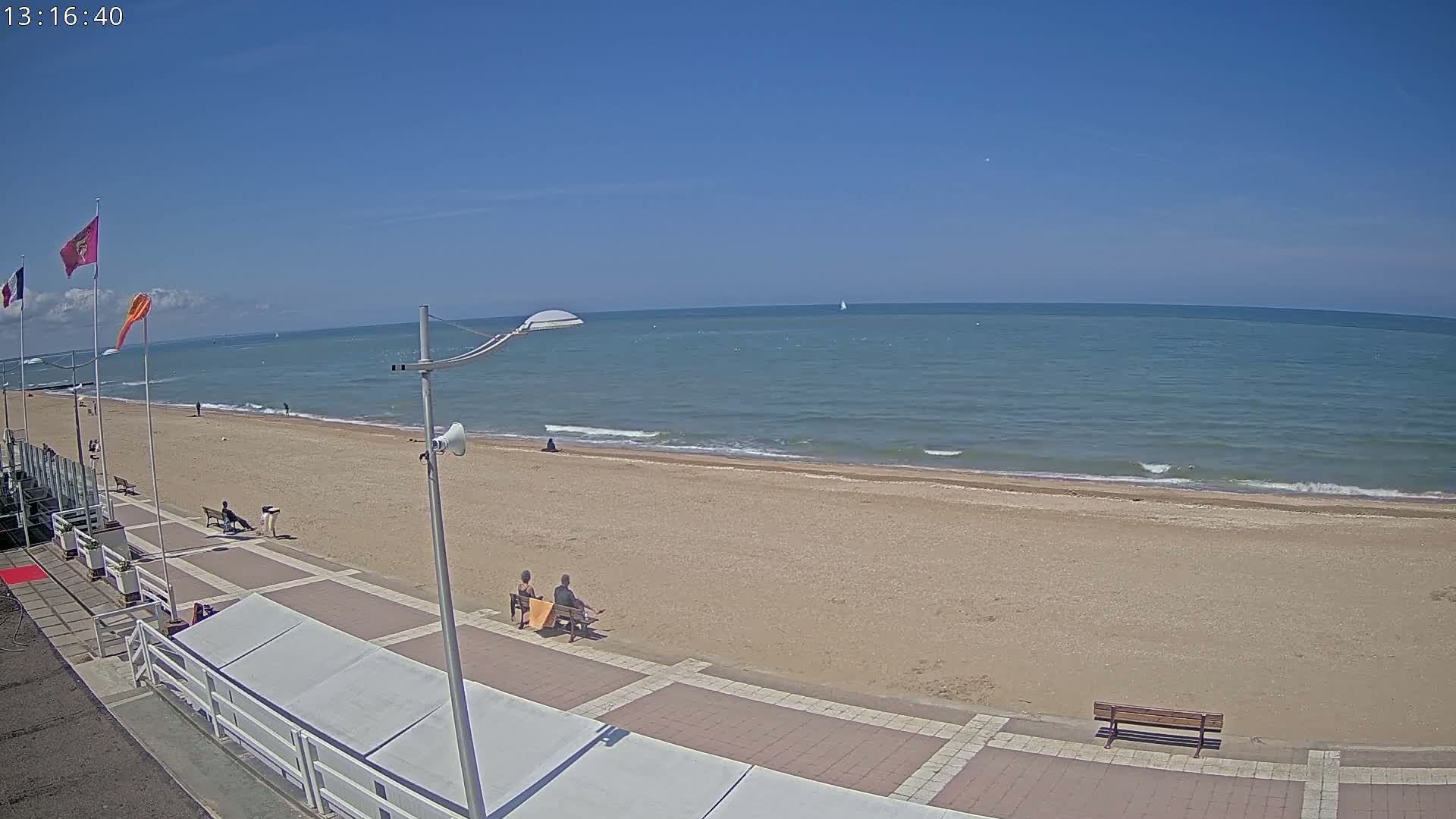 A sandy beach with a paved walkway and several people sitting on benches, under a clear blue sky on a sunny day, with calm ocean waves in the background.