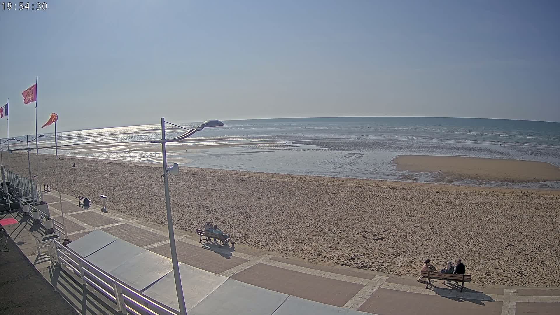 A sandy beach with calm ocean waves under a clear blue sky, featuring several people sitting on benches near a paved walkway.
