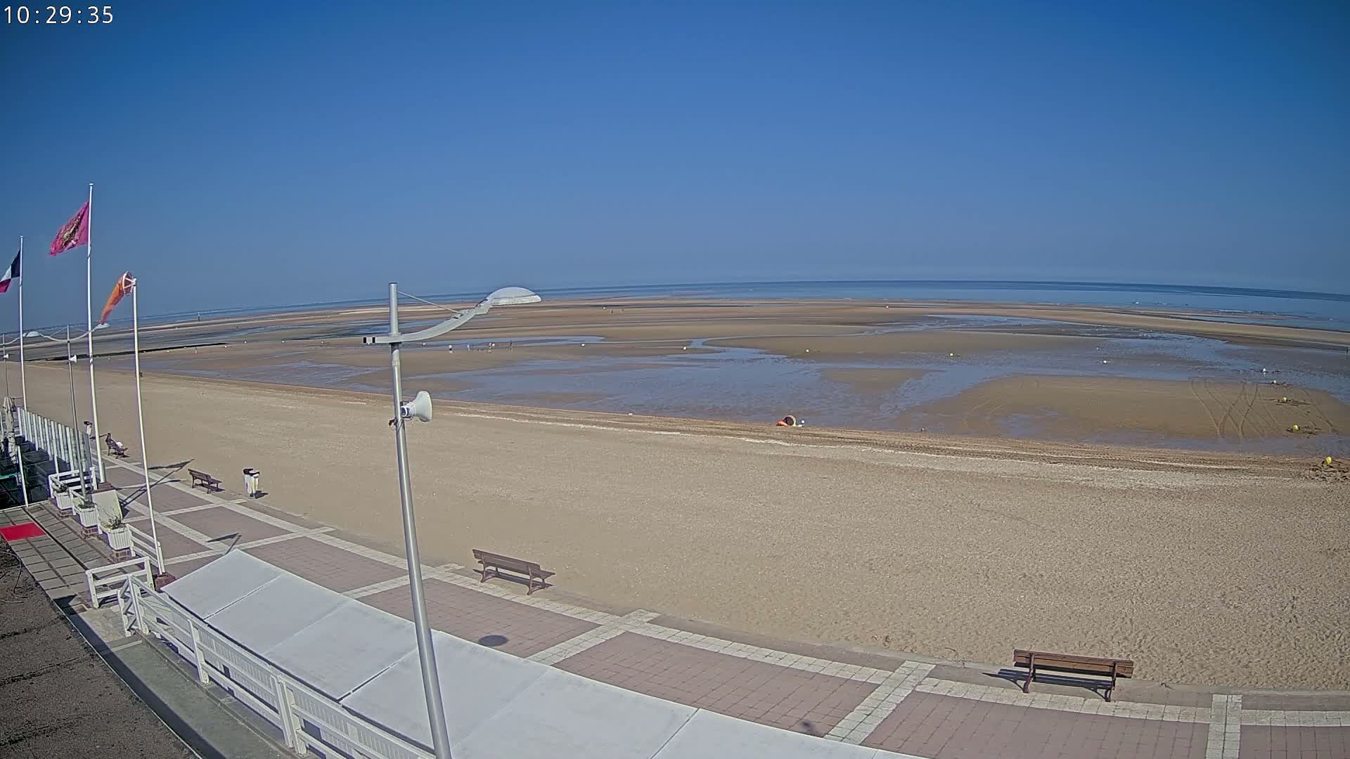 A wide shot of a sandy beach on a sunny day shows a mostly empty beach with a paved boardwalk and a few benches.