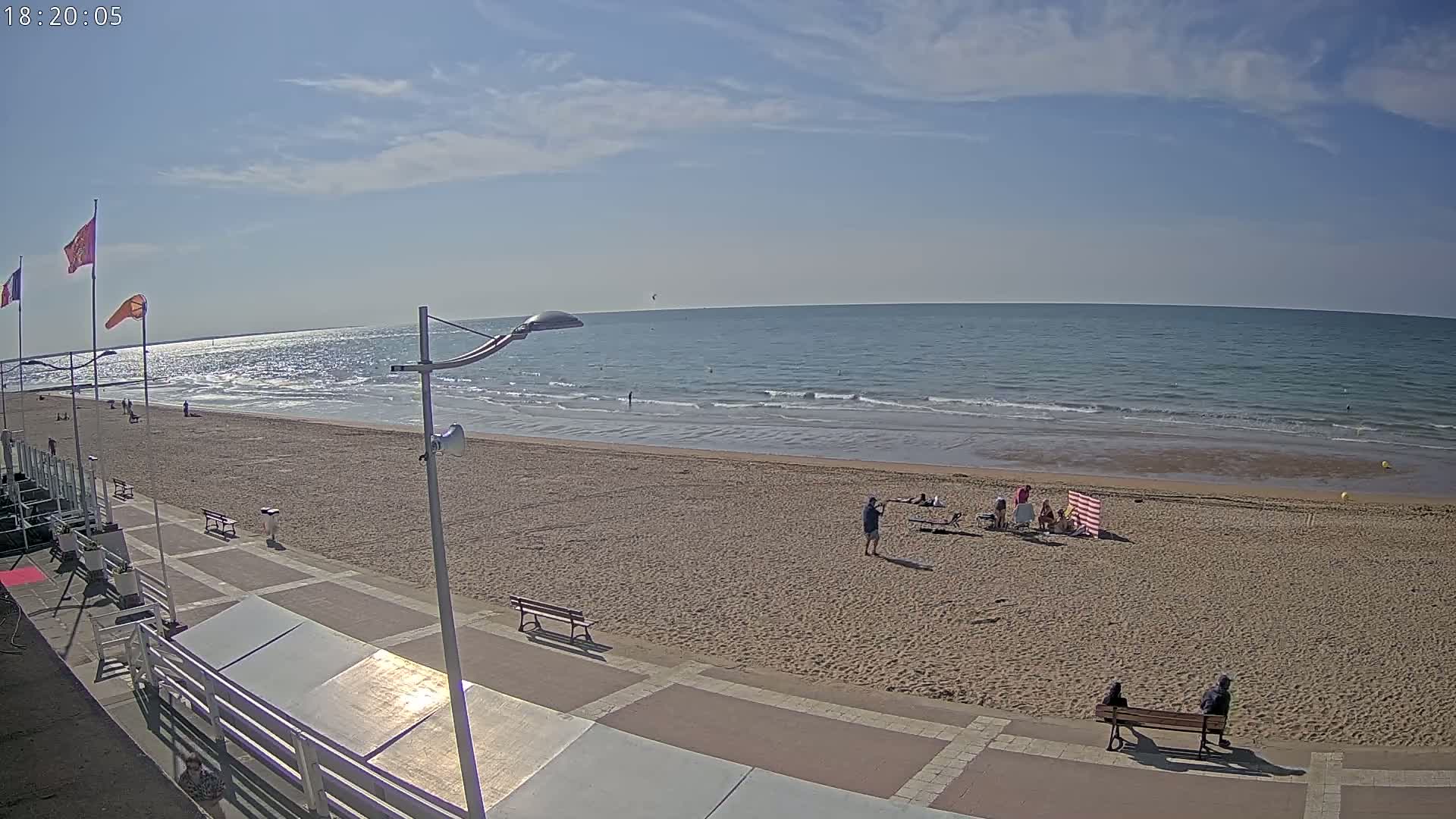 A sandy beach with a few people and benches, a boardwalk, and a calm ocean under a mostly sunny sky.