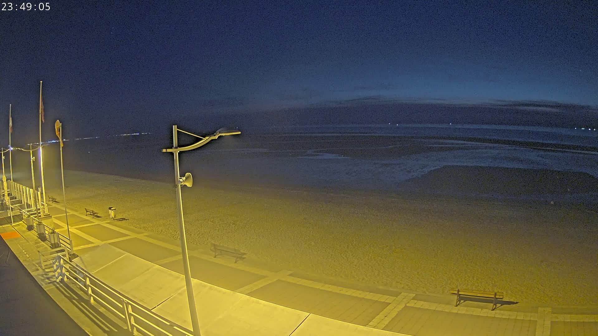 A nighttime view of a beach with a boardwalk, showing a mostly empty sandy shore under a dark blue sky.