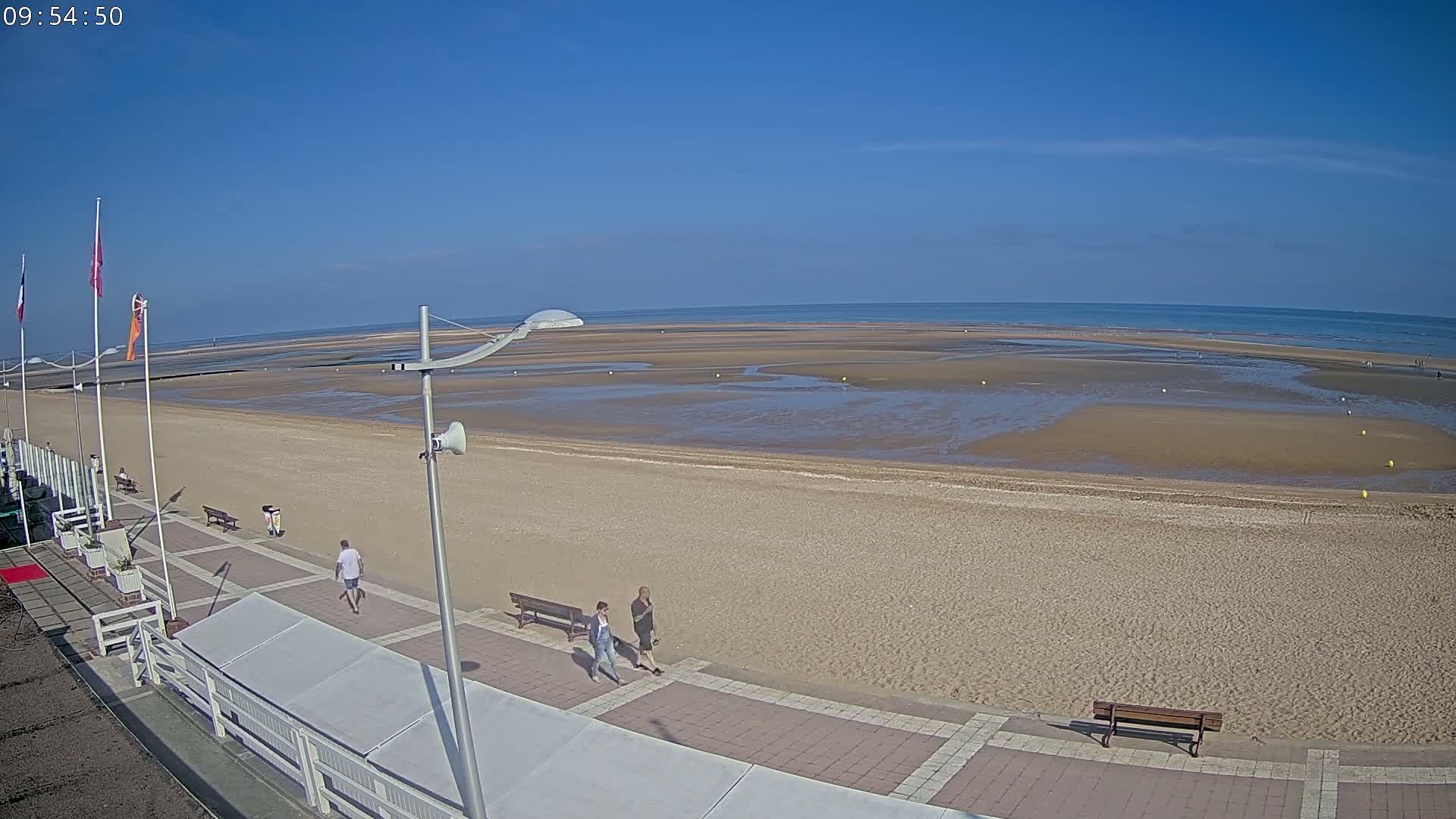 A wide sandy beach stretches to a calm ocean under a clear blue sky, with a paved walkway and several people strolling along the beachfront.