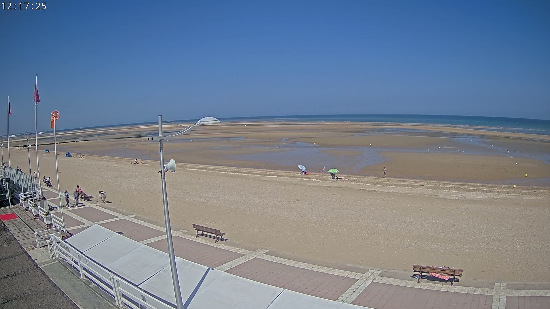 A sandy beach with a few people and umbrellas under a clear blue sky stretches to a calm ocean.