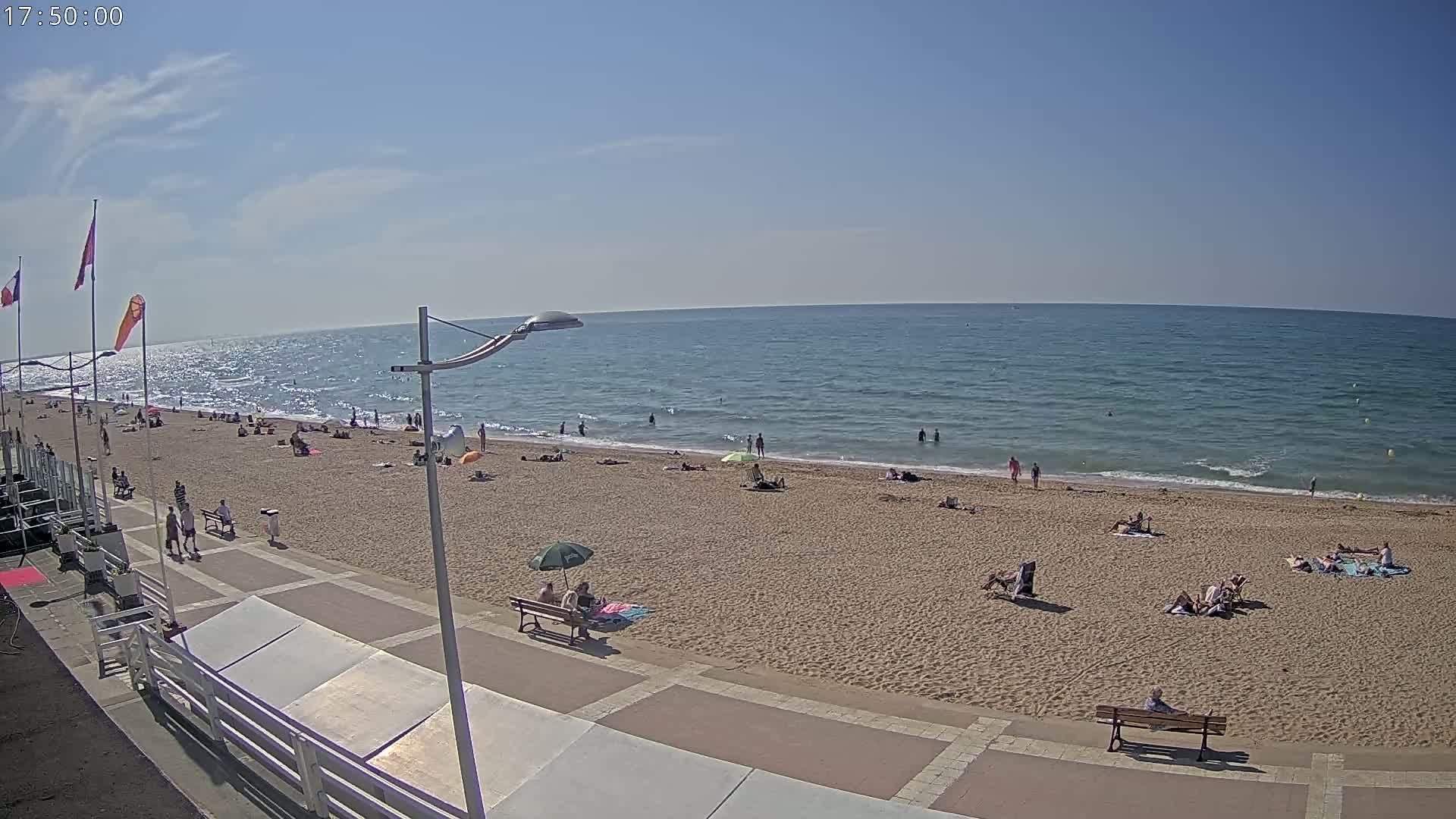 A sandy beach with scattered sunbathers and a calm, blue ocean under a mostly sunny sky.